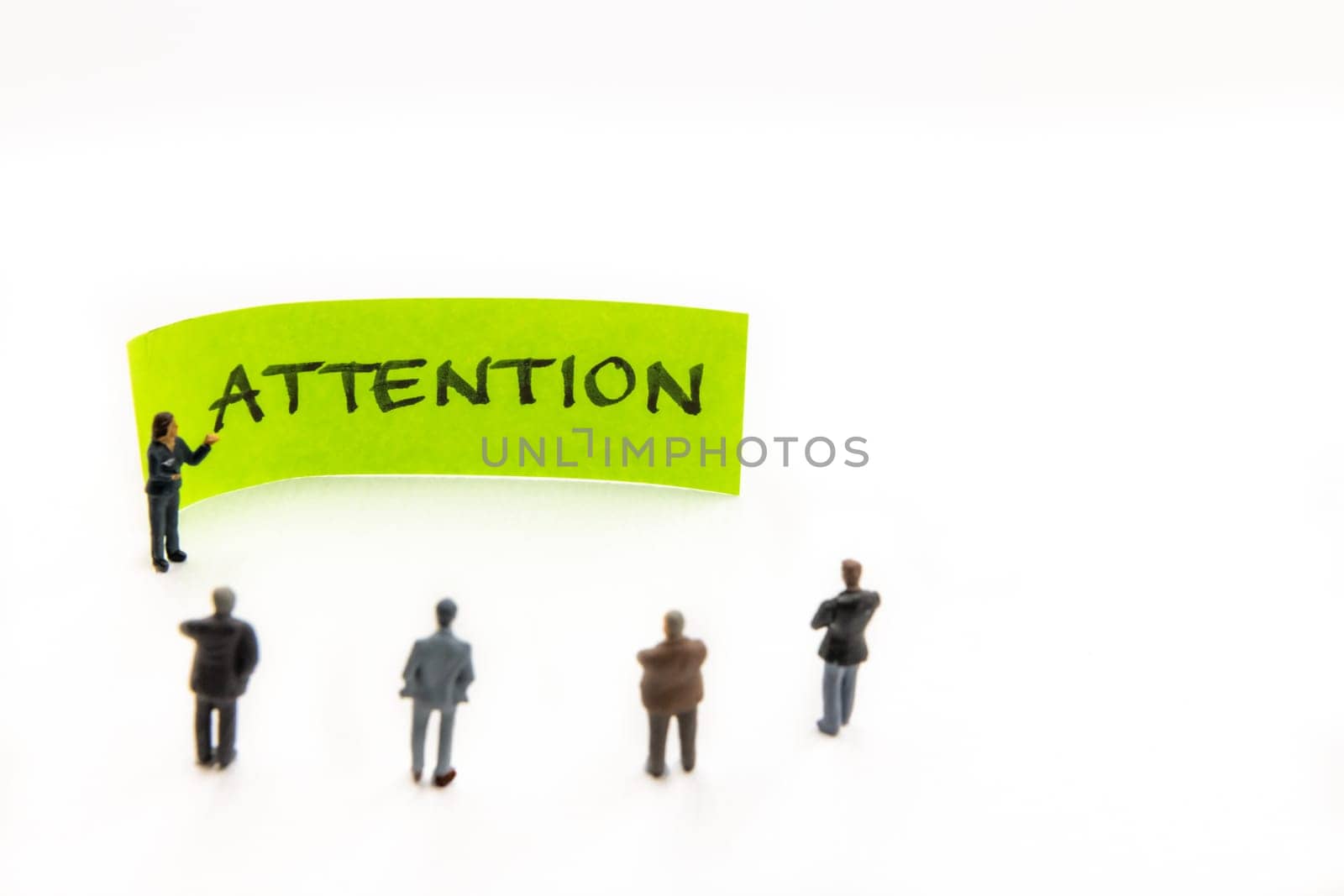 Strategy meeting with miniature figurines posed as business people standing around post-it note with Attention handwritten message on white background, minimalist abstract concept with focus on text