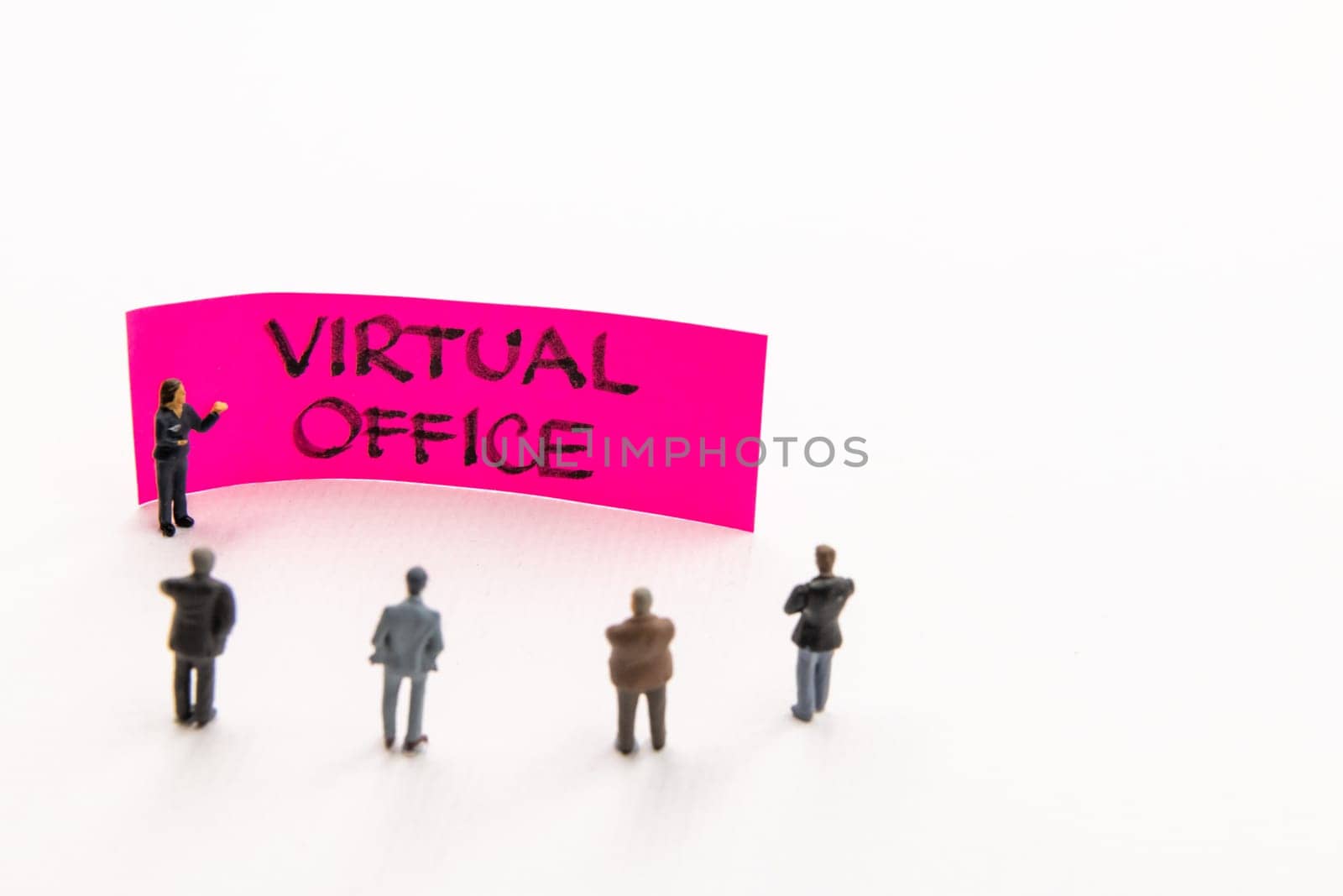 Strategy meeting with miniature figurines posed as business people standing around post-it note with Virtual Office handwritten message on white background, minimalist abstract concept with focus on text