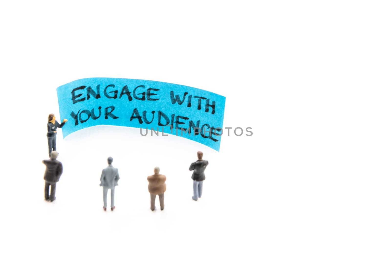 Meeting with miniature figurines posed as business people standing around post-it note with Engage With Your Audience handwritten message in background, minimalist abstract concept with focus on text