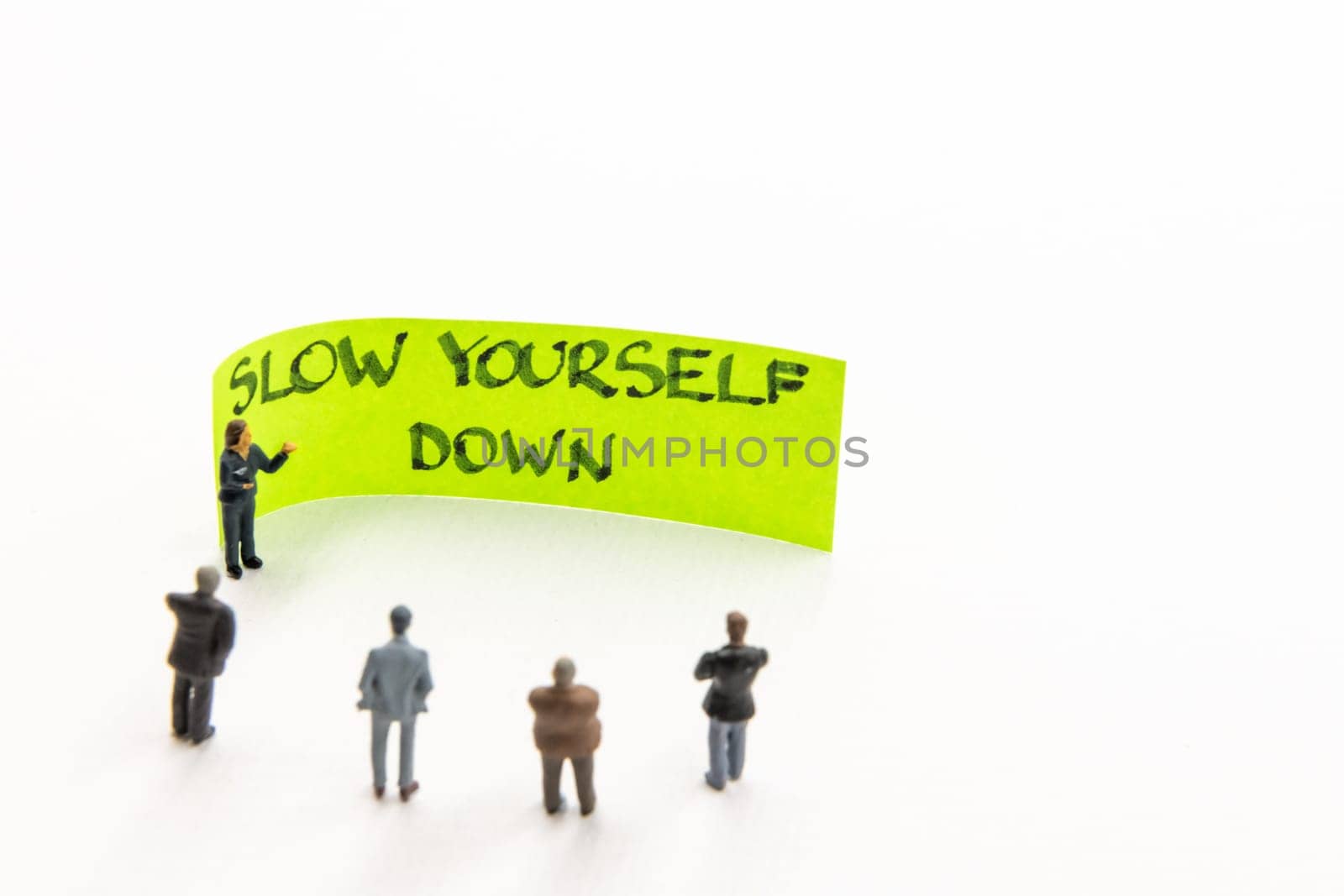 Meeting with miniature figurines posed as business people standing around post-it note with Slow Yourself Down handwritten message on white background, minimalist abstract concept with focus on text