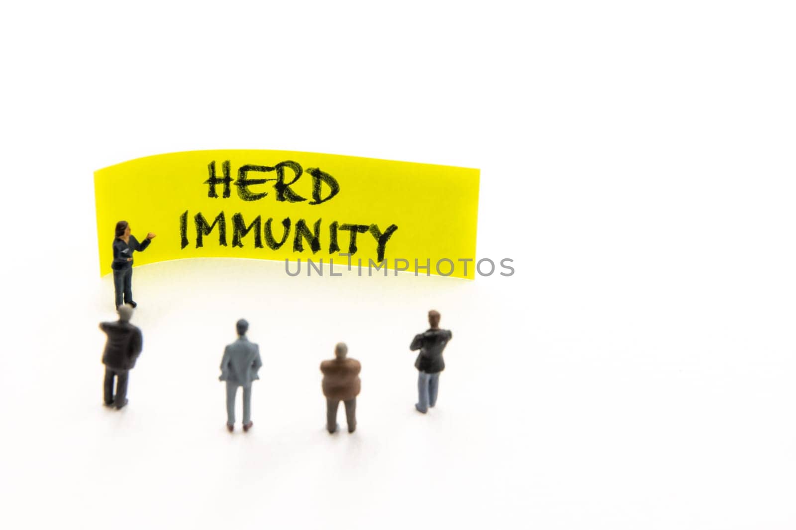 Meeting with miniature figurines posed as business people standing around post-it note with Herd Immunity handwritten message in background, minimalist abstract concept with focus on text