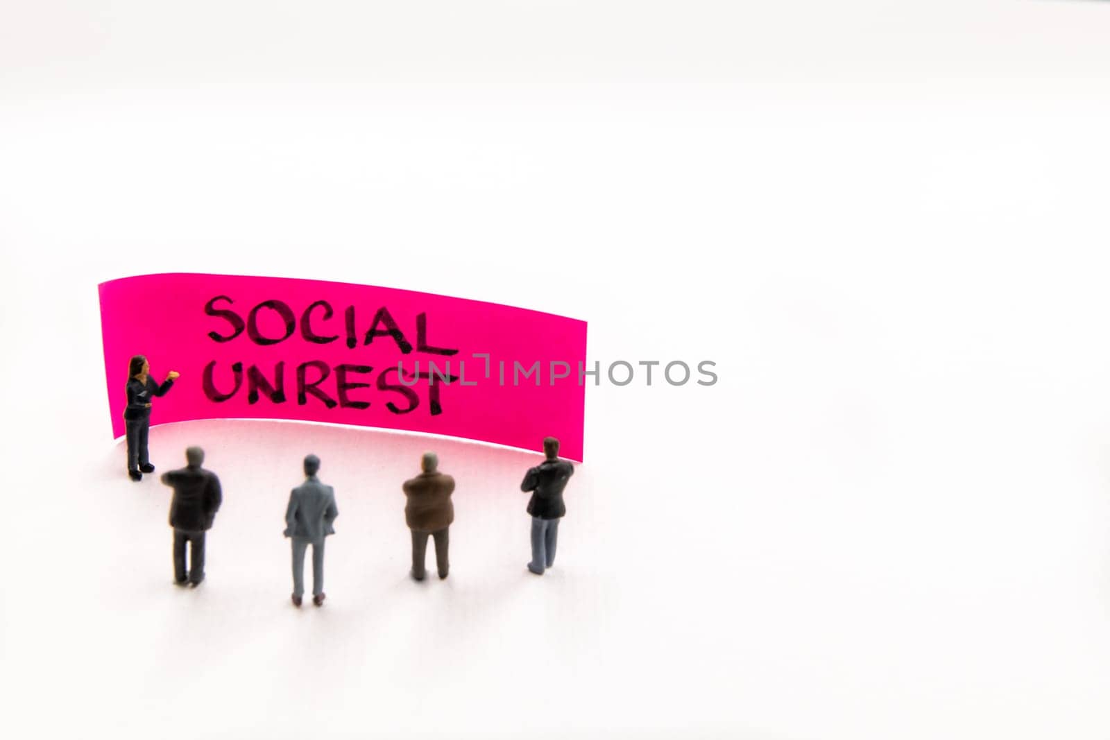 Meeting with miniature figurines posed as business people standing around post-it note with Social Unrest handwritten message in background, minimalist abstract concept with focus on text