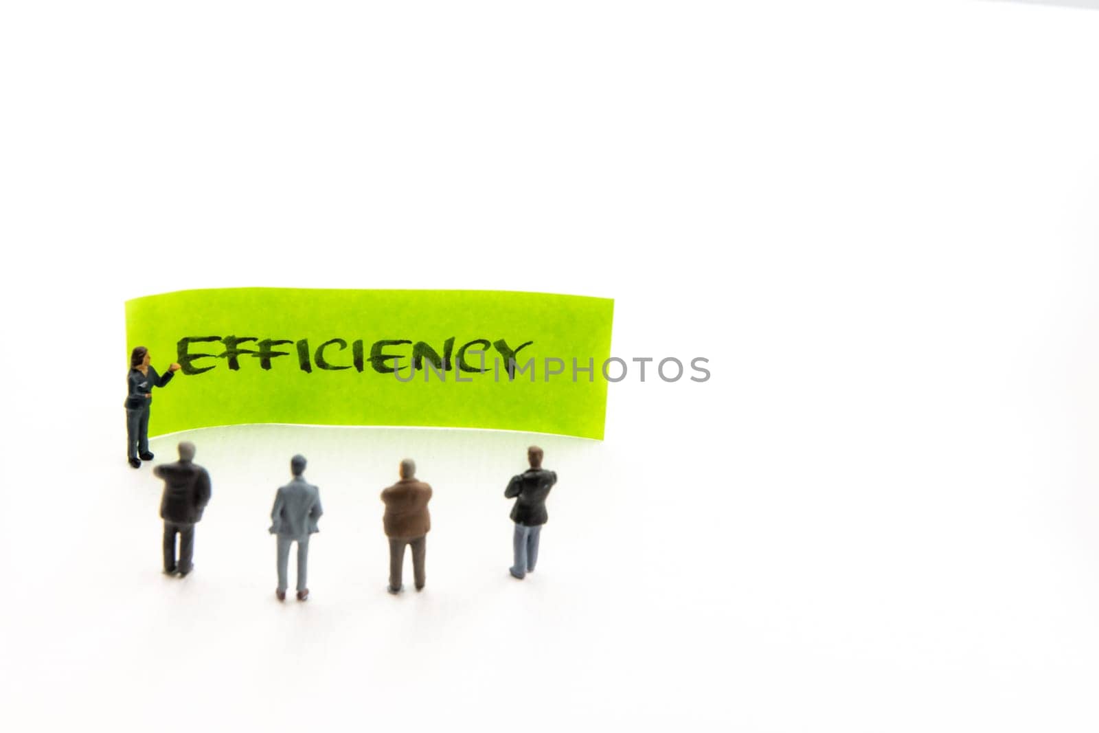 Meeting with miniature figurines posed as business people standing around post-it note with Efficiency handwritten message in background, minimalist abstract concept with focus on text