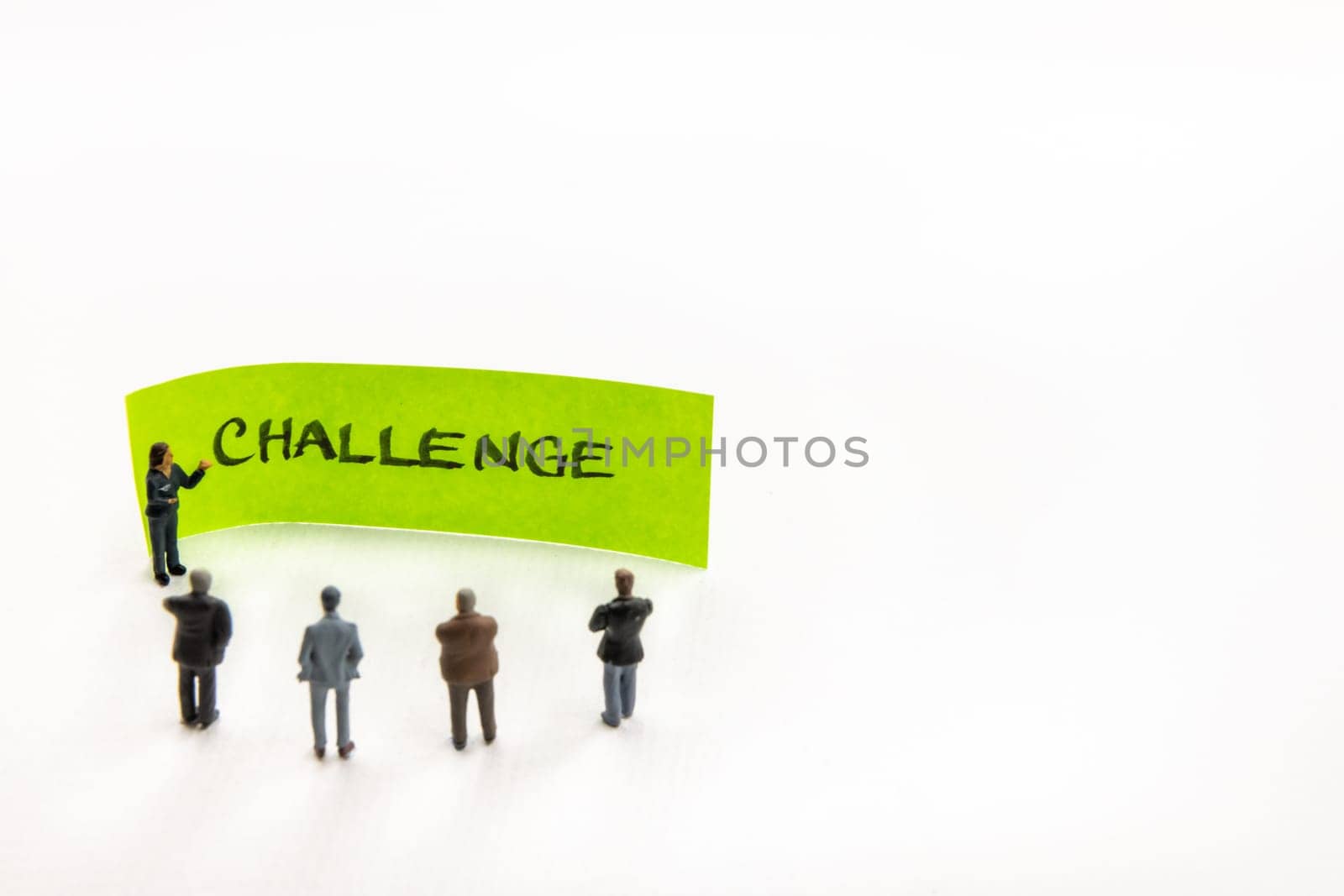 Meeting with miniature figurines posed as business people standing around post-it note with Challenge handwritten message in background, minimalist abstract concept with focus on text