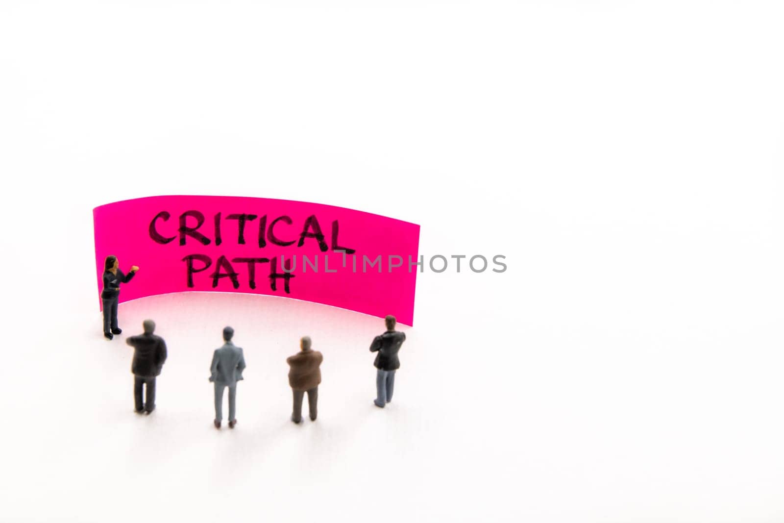 Meeting with miniature figurines posed as business people standing around post-it note with Critical Path handwritten message in background, minimalist abstract concept with focus on text