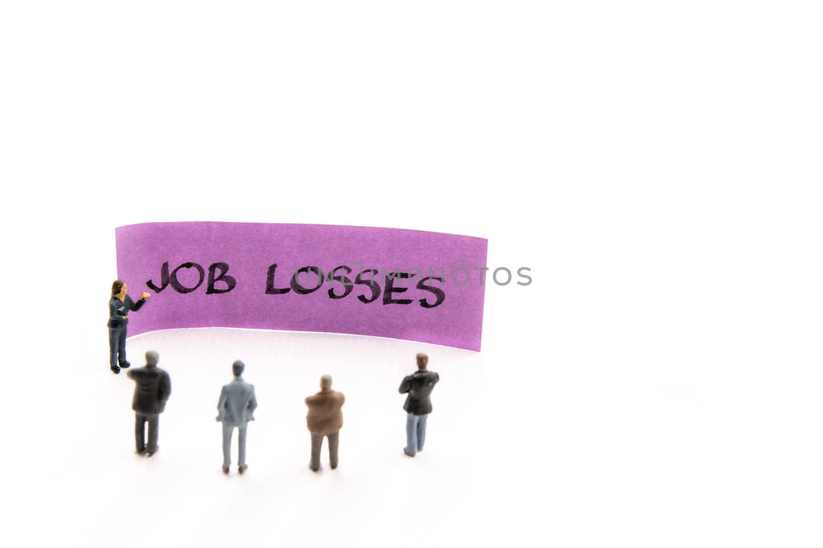 Meeting with miniature figurines posed as business people standing around post-it note with Job Losses handwritten message in background, minimalist abstract concept with focus on text