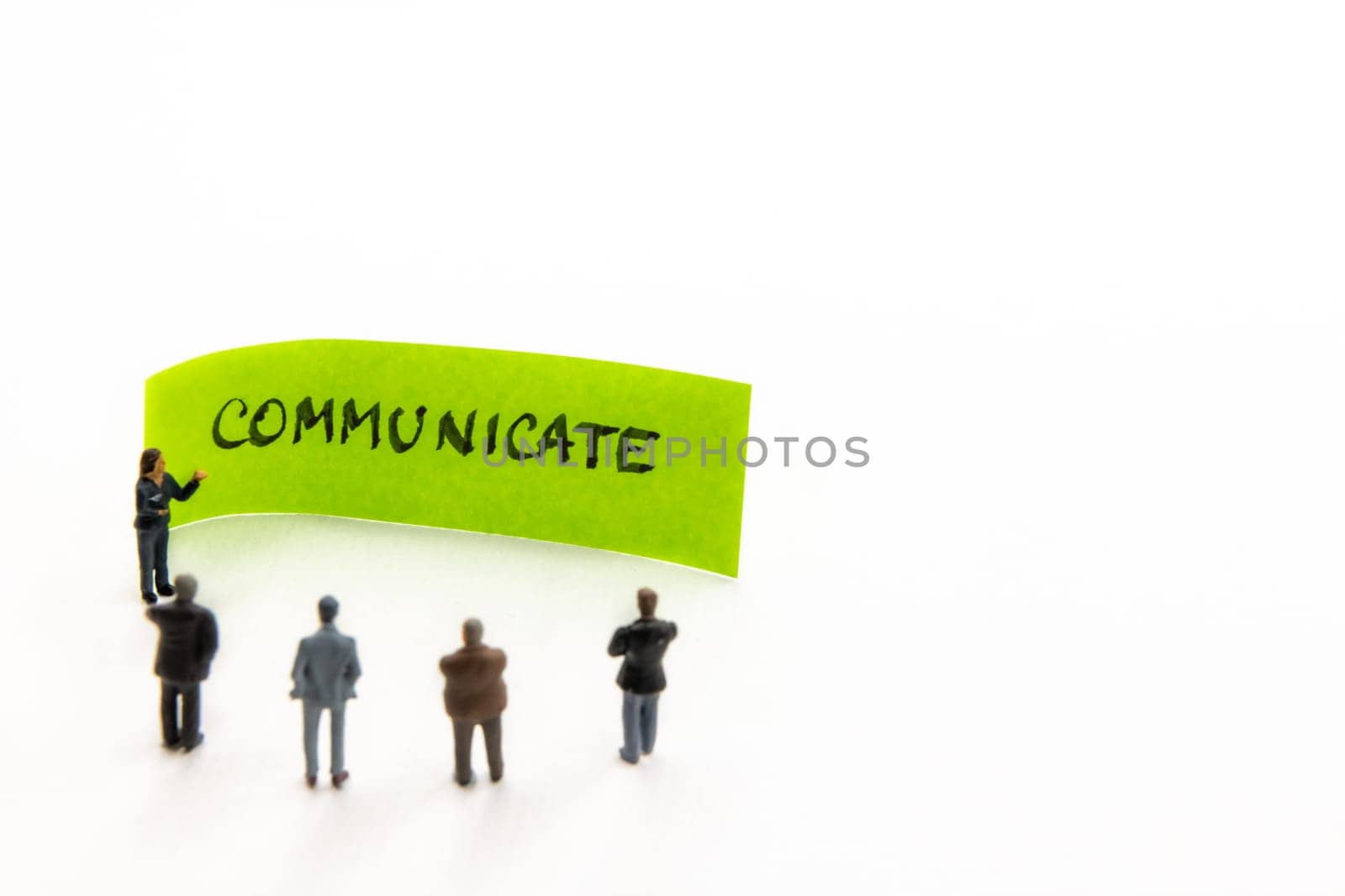 Meeting with miniature figurines posed as business people standing around post-it note with Communicate handwritten message in background, minimalist abstract concept with focus on text