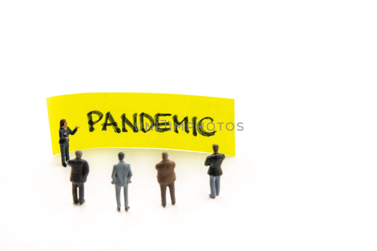 Meeting with miniature figurines posed as business people standing around post-it note with Pandemic handwritten message in background, minimalist abstract concept with focus on text