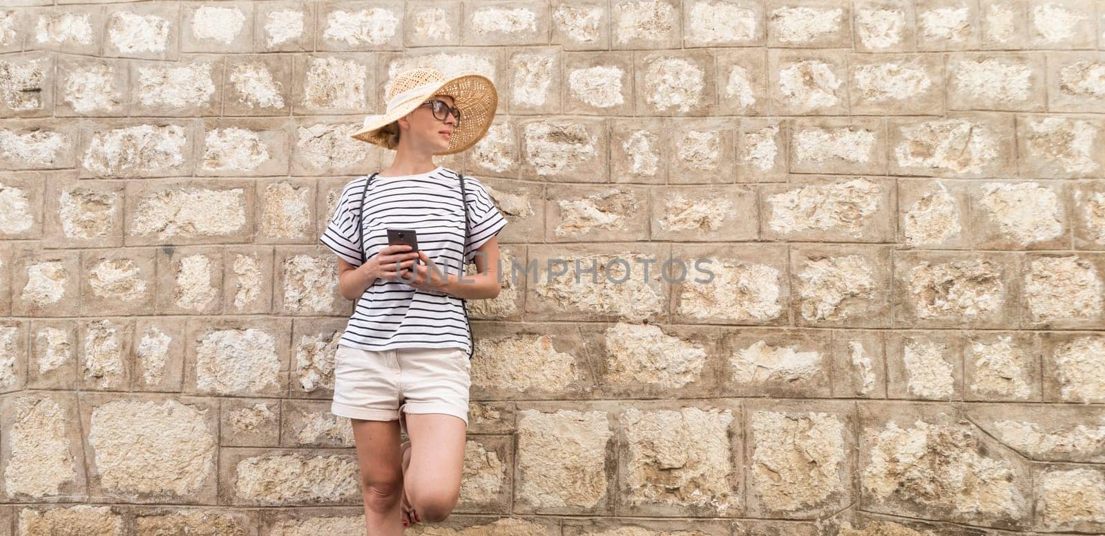 Beautiful young female tourist woman standing in front of old textured stone wall at old Mediterranean town, smiling, holding, smart phone to network on vacationes. Copy space.