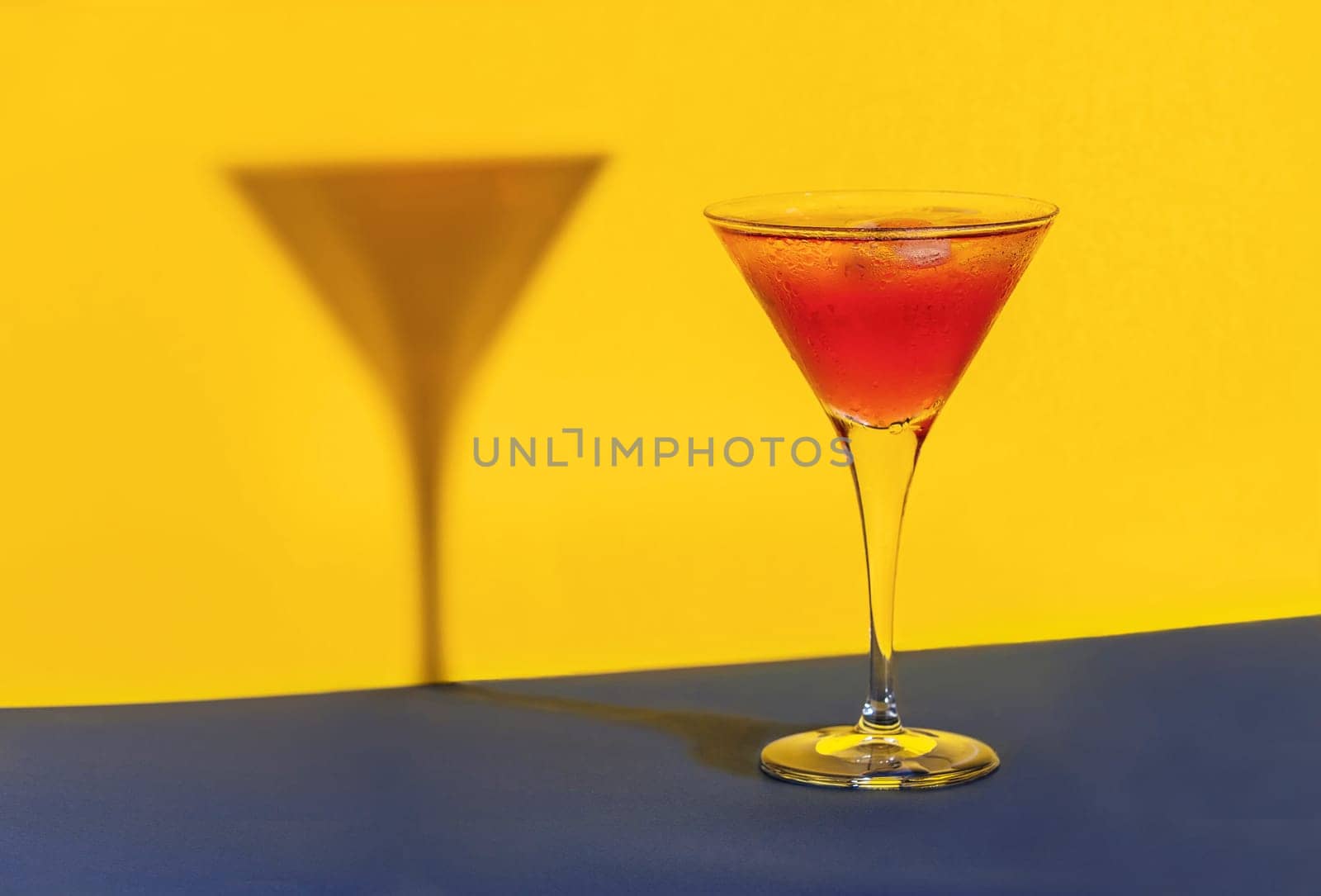 Red cocktail in a glass shaping its shadow on a blue and yellow background