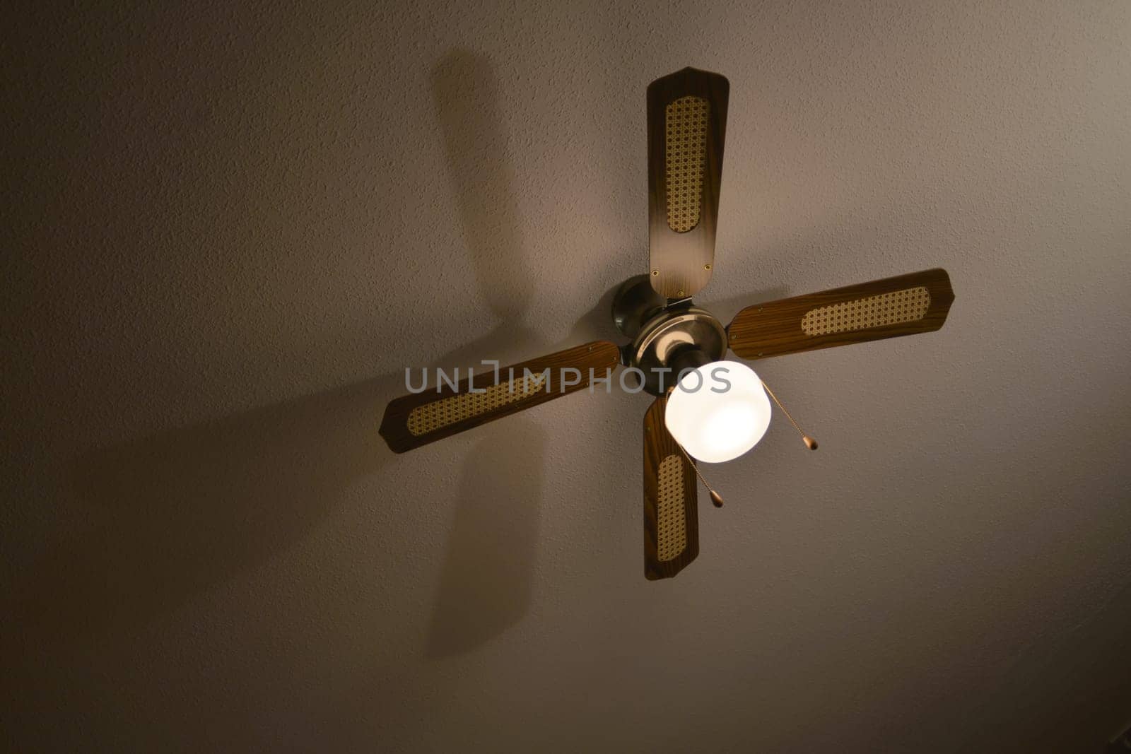Mediterranean brown wood and material ceiling fan casts a long shadow on a plaster ceiling.