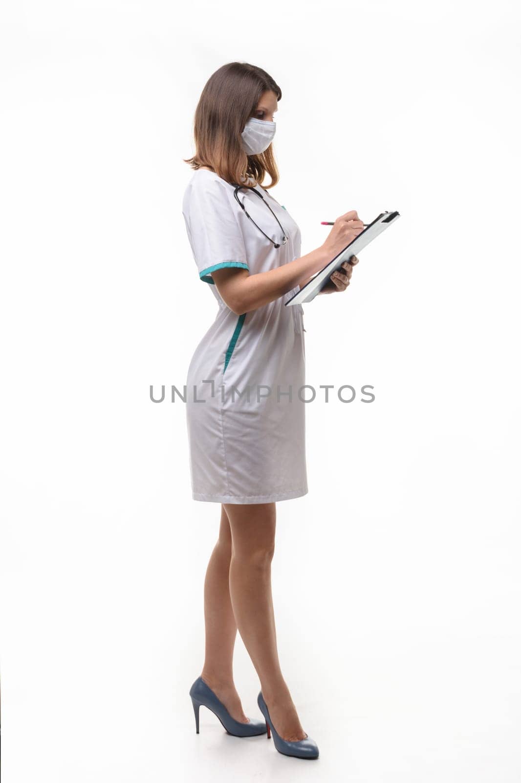 A girl in medical clothes and a mask stands sideways and writes in a tablet information on a white background