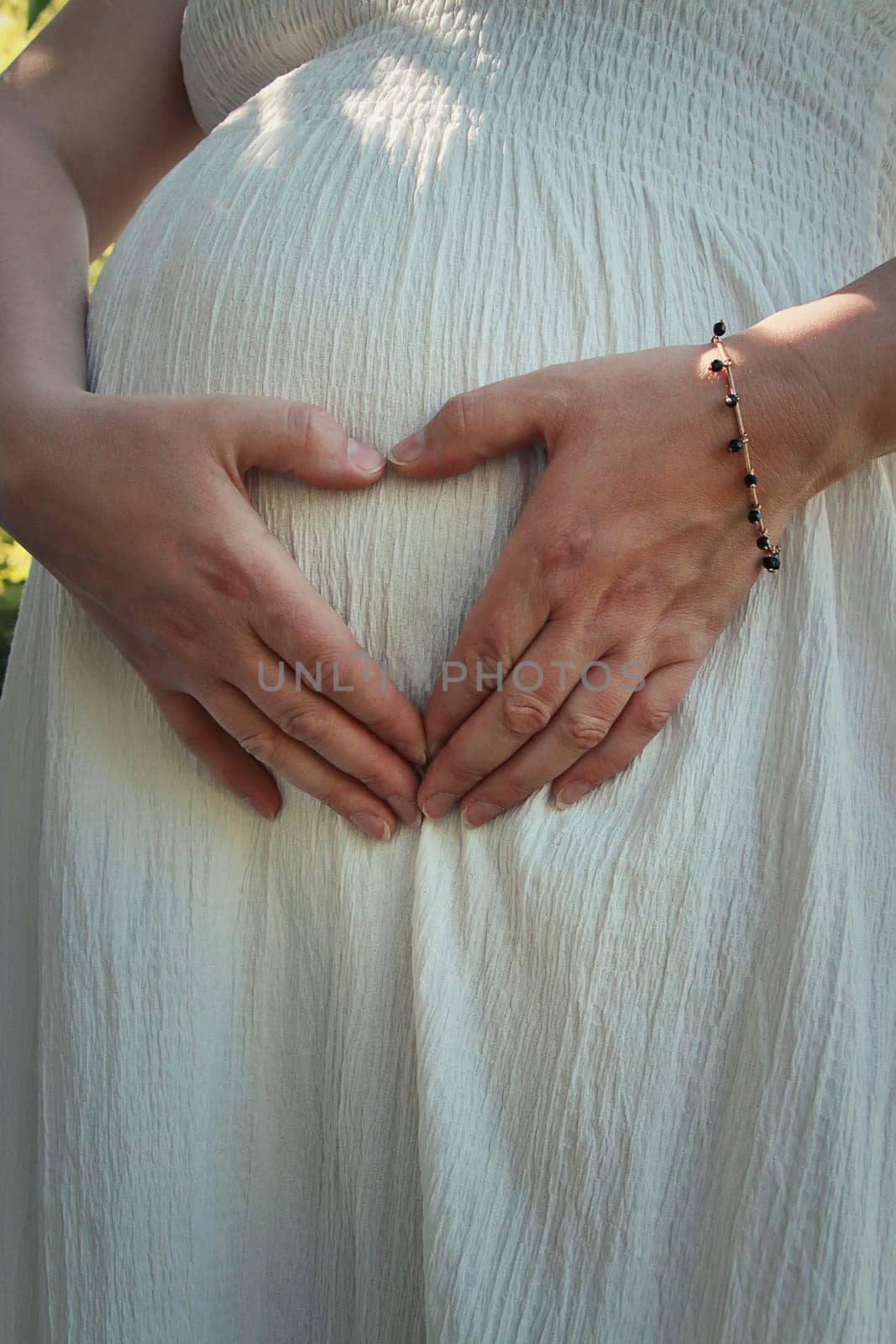 Closeup cute composition with a pregnant young woman wearing white dress makes a heart shaped symbol with her hands on her belly. Happy mother waiting for a baby. Family love concept, motherhood.