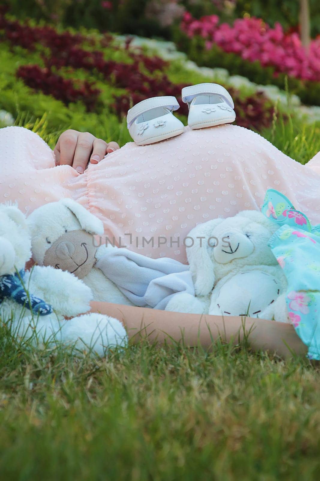 Cute composition with a pregnant young woman laying outdoors on the grass in the park surrounded by multiple baby stuffed toys and newborn shoes on her belly. Happy mother and family waiting for child