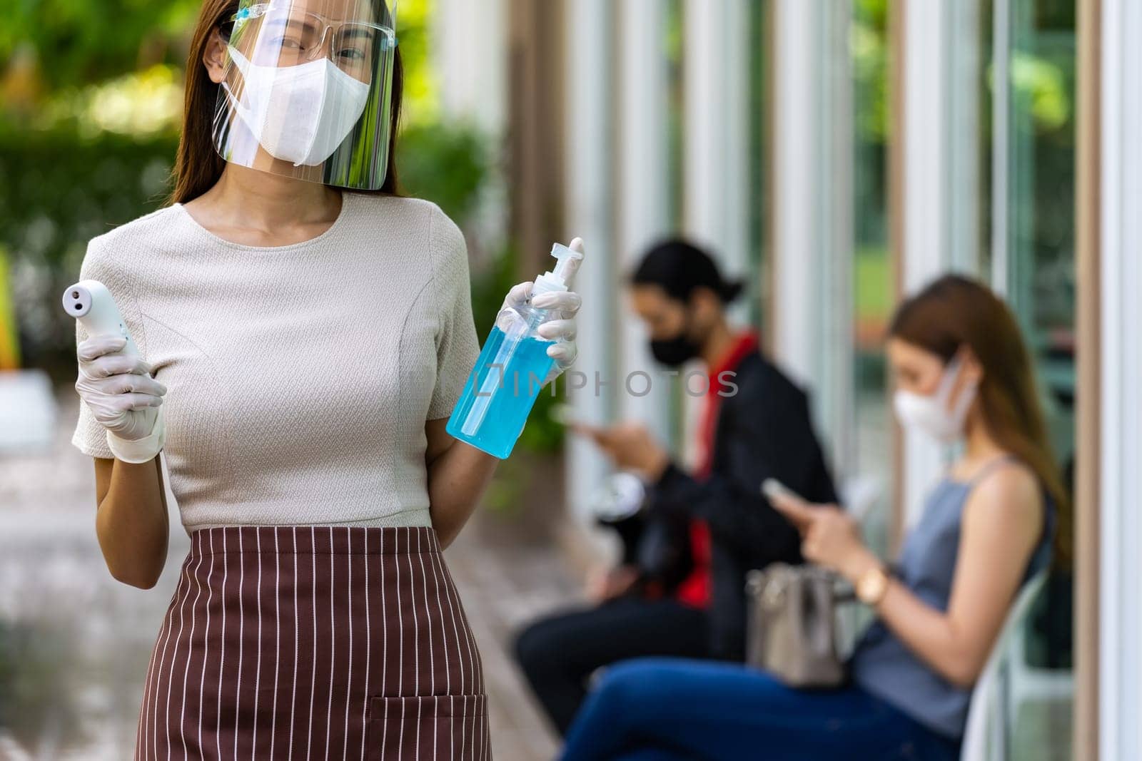 Portrait attractive asian waitress wear face mask and shield hold thermometer and alcohol gel sanitizer with background of social distancing customer waiting in queue. New normal restaurant concept.