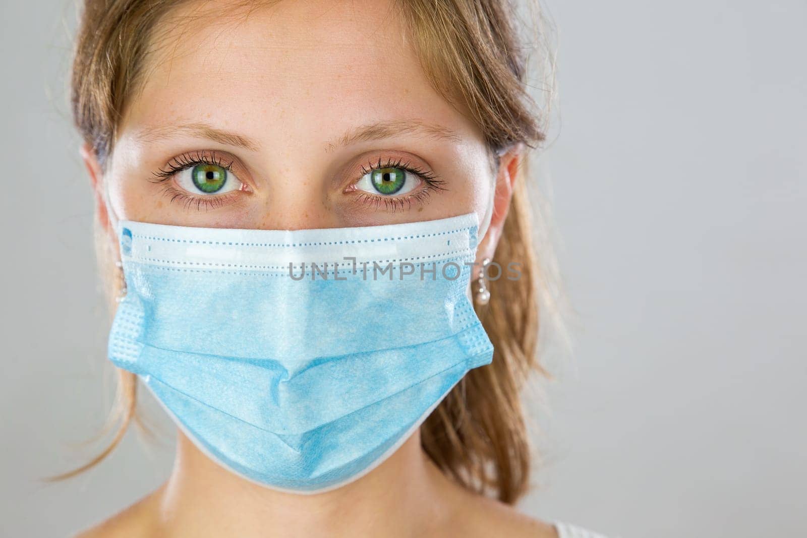 Young woman with green eyes wearing blue surgical face mask and facing camera in studio. Blond lady staring from front with protection over face to prevent infection.