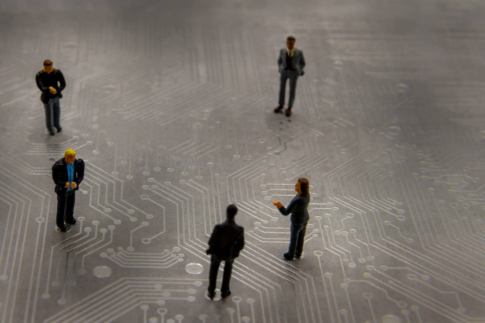 Miniature figurines posed as business people in a meeting over abstract futuristic circuit board, future business concept
