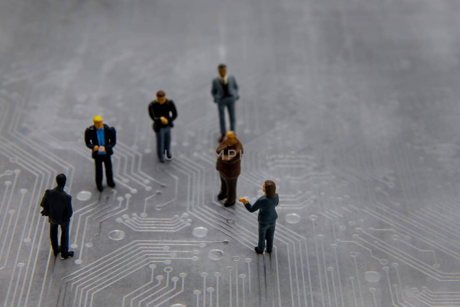 Miniature figurines posed as business people in a meeting over abstract futuristic circuit board, future business concept