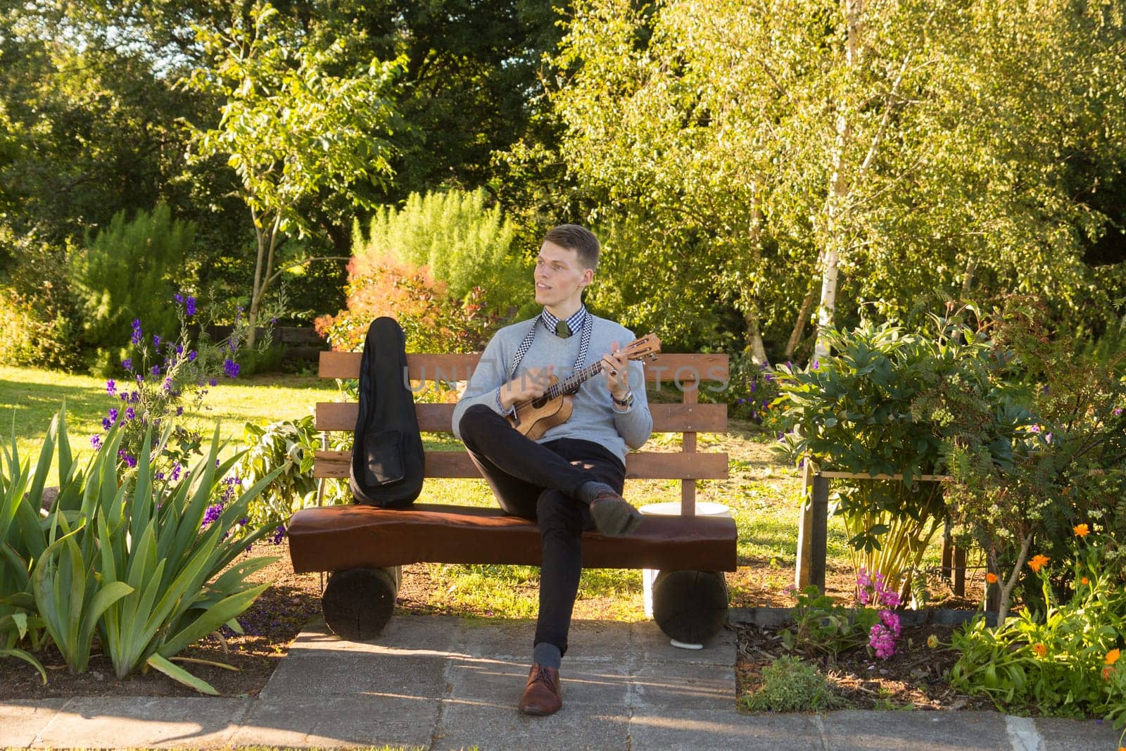 Young man sitting on bench in the park and playing ukulele.