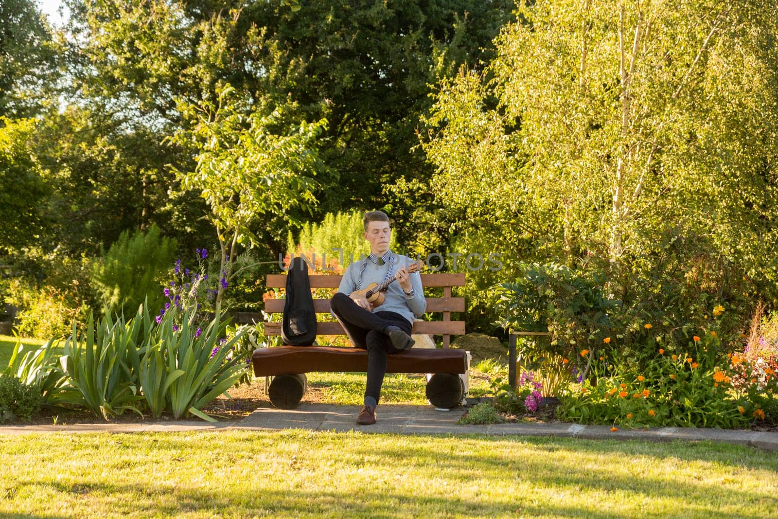 Young man sitting on bench in the park and playing ukulele.