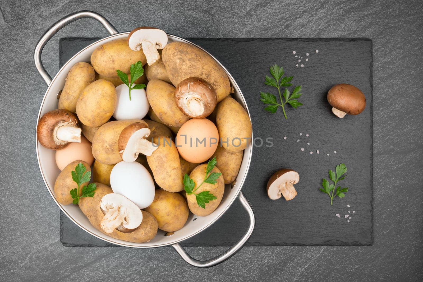 Raw potatoes, eggs, halved and whole brown mushrooms, coarse salt grains and parsley leaves in a white enamel sieve on a slate slap. Flat lay.