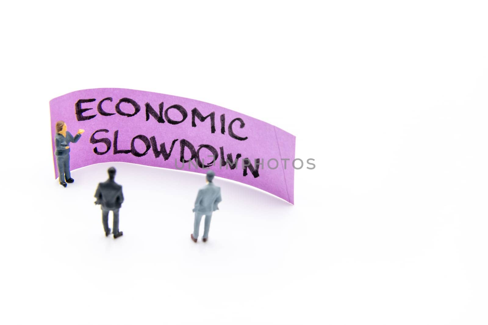 Presentation meeting with miniature figurines posed as business people standing in front of post-it note with Economic Slowdown handwritten message in background, copy space on the right