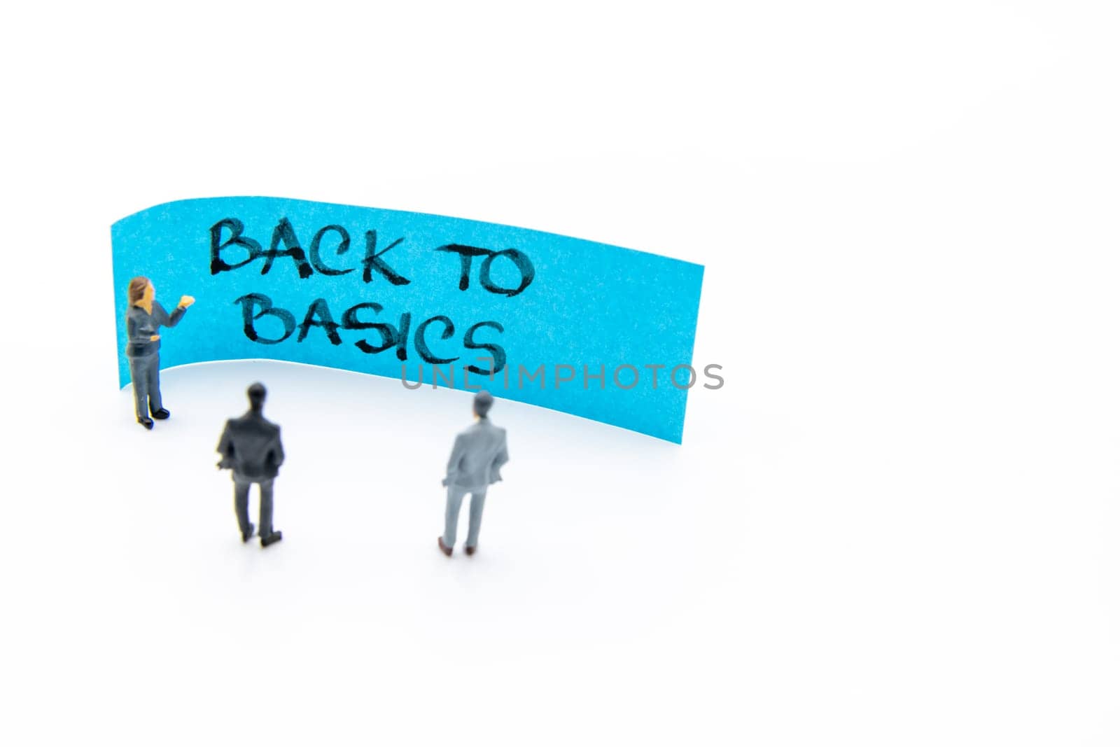 Presentation meeting with miniature figurines posed as business people standing in front of post-it note with Back To Basics handwritten message in background, copy space on the right