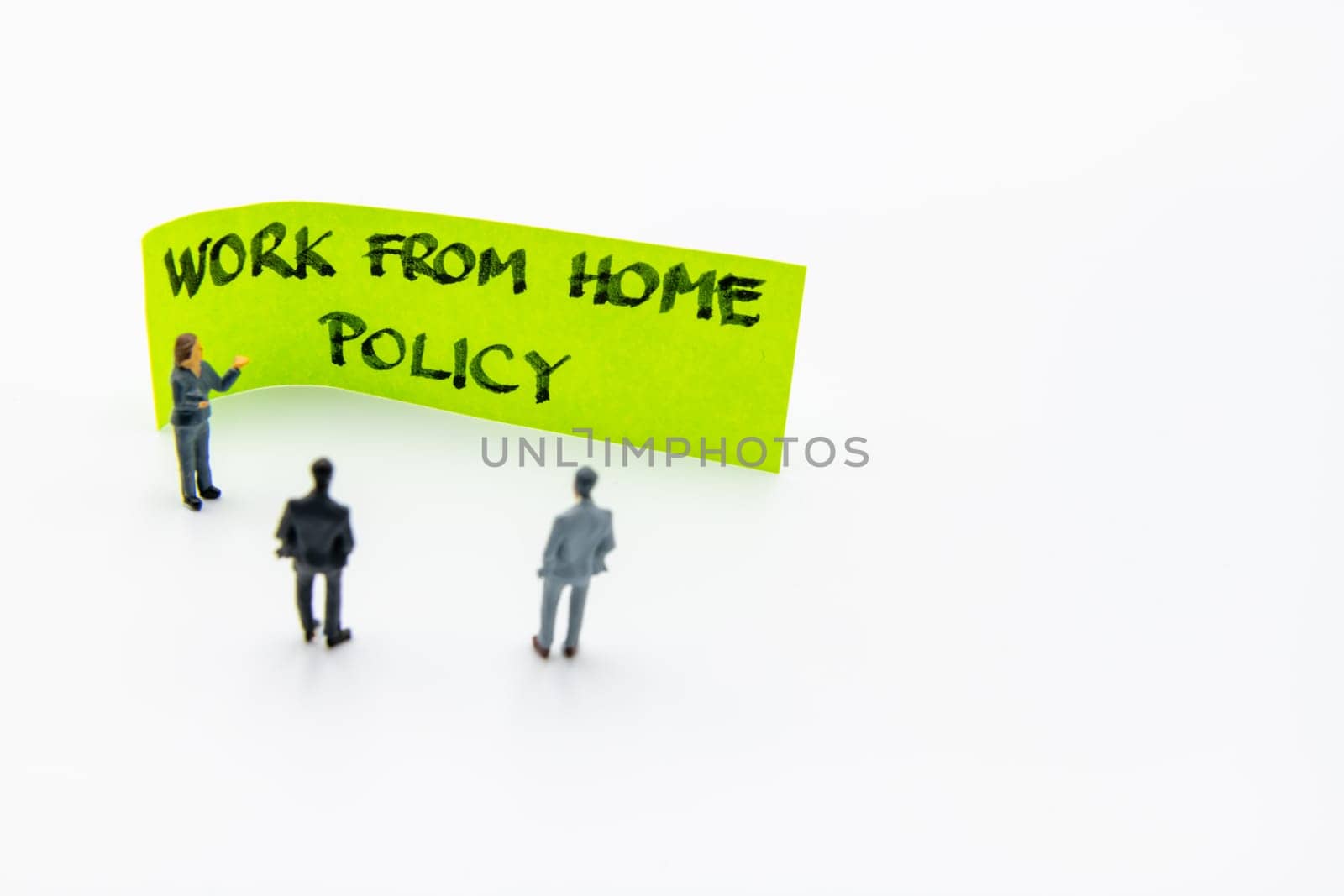 Presentation meeting with miniature figurines posed as business people standing in front of post-it note with Work From Home Policy handwritten message in background, copy space on the right