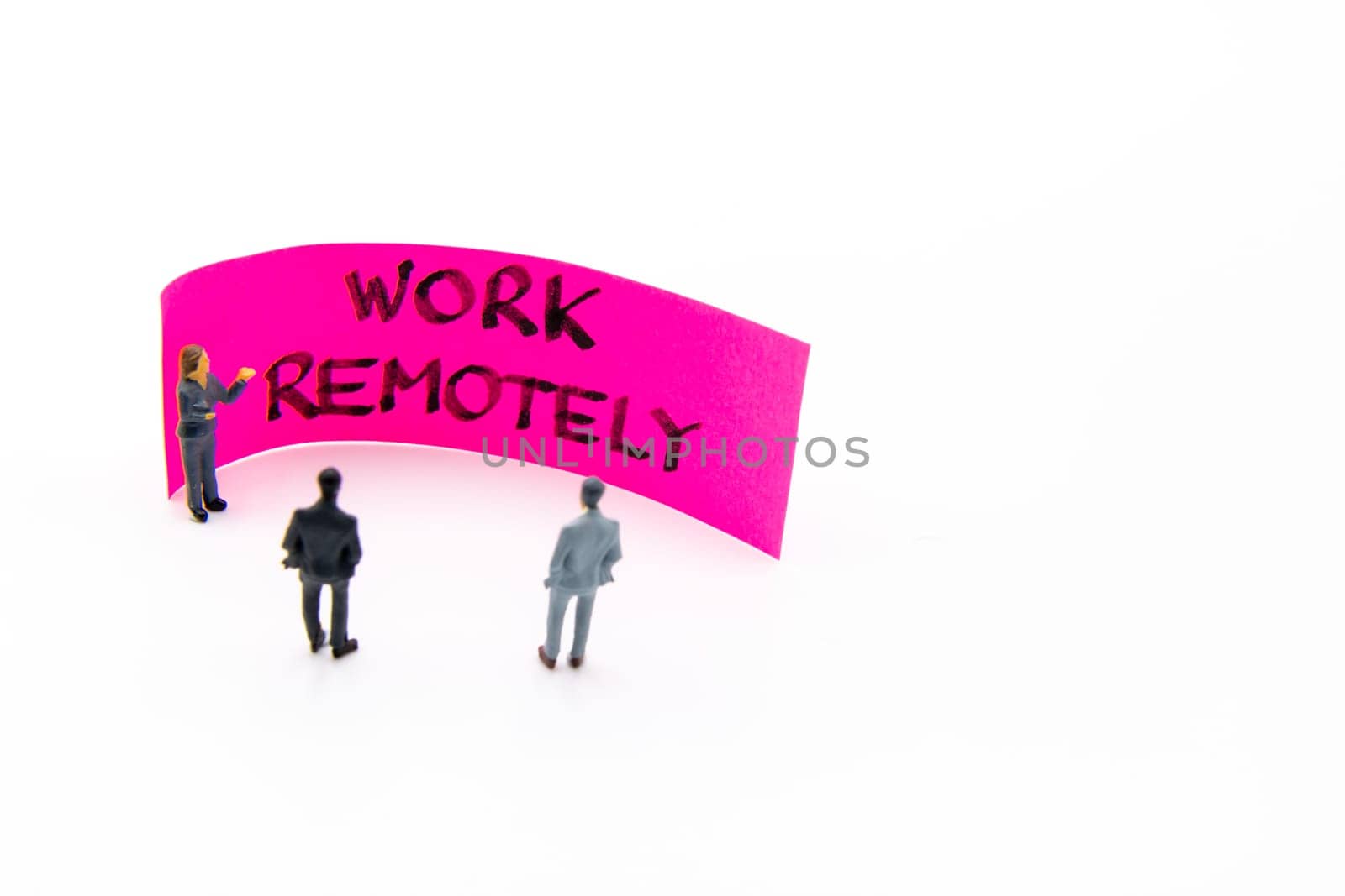 Presentation meeting with miniature figurines posed as business people standing in front of post-it note with Work Remotely handwritten message in background, copy space on the right