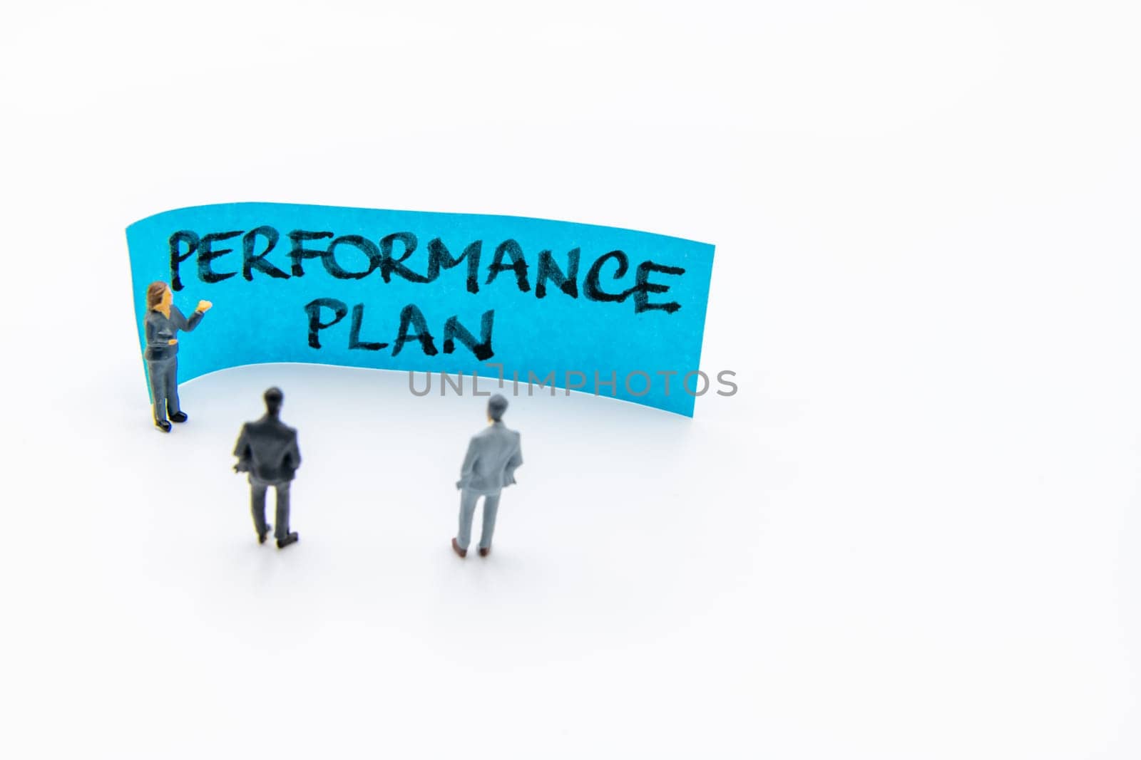 Presentation meeting with miniature figurines posed as business people standing in front of post-it note with Performance Plan handwritten message in background, copy space on the right