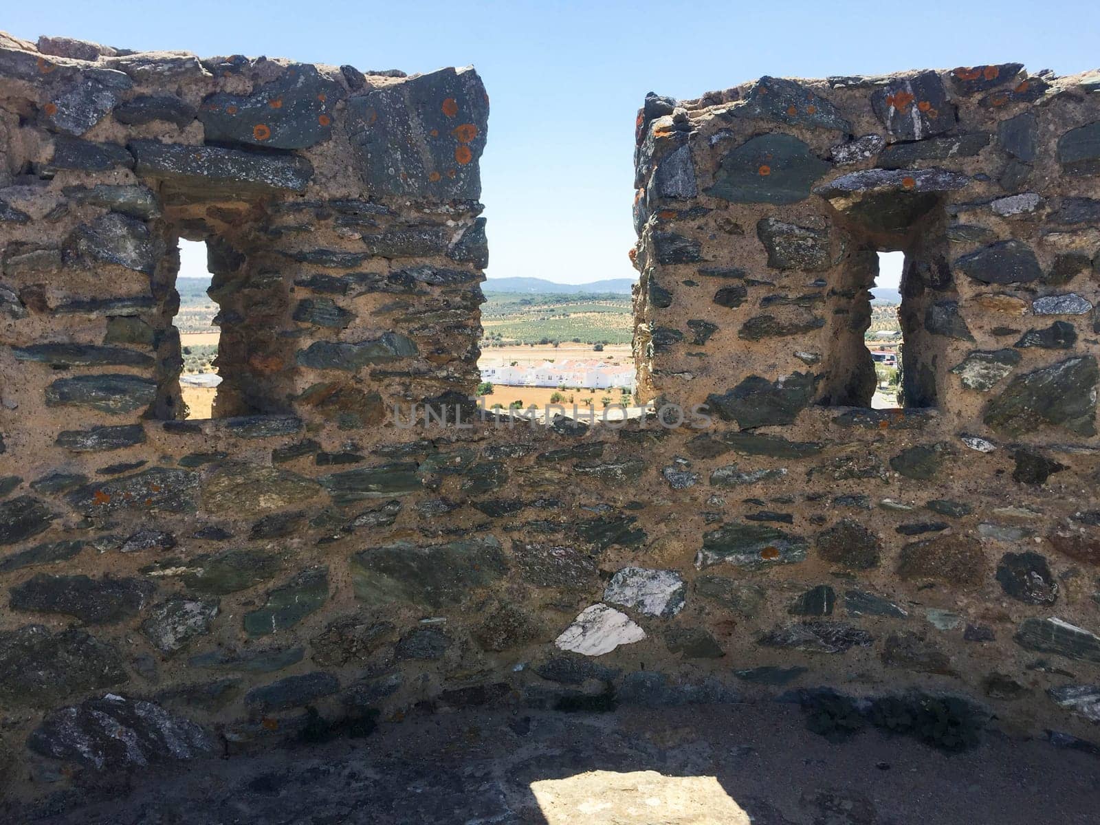 Ancient stone battlements and loopholes with view of countryside and distant horizon under blue sky in natural light. Full frame of rough stone texture and gentle nature with copy space.
