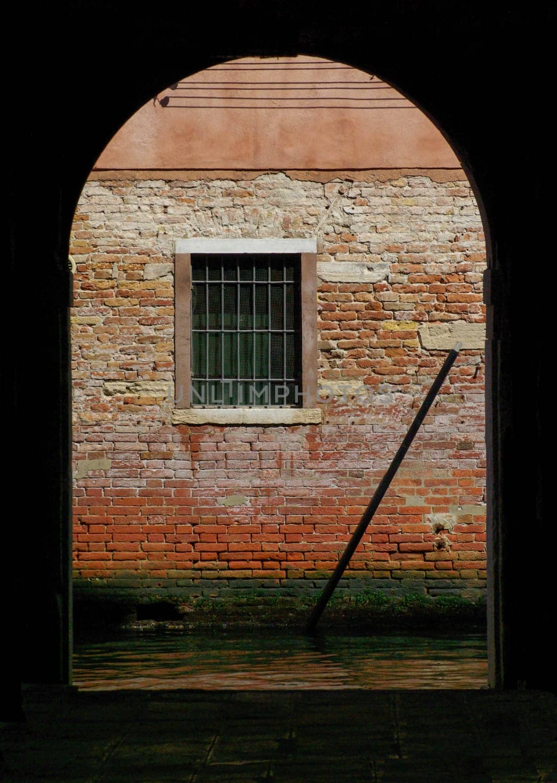 Dark interior opens through archway to ancient red brick wall and window with bars. Beautiful green and red colors and texture.