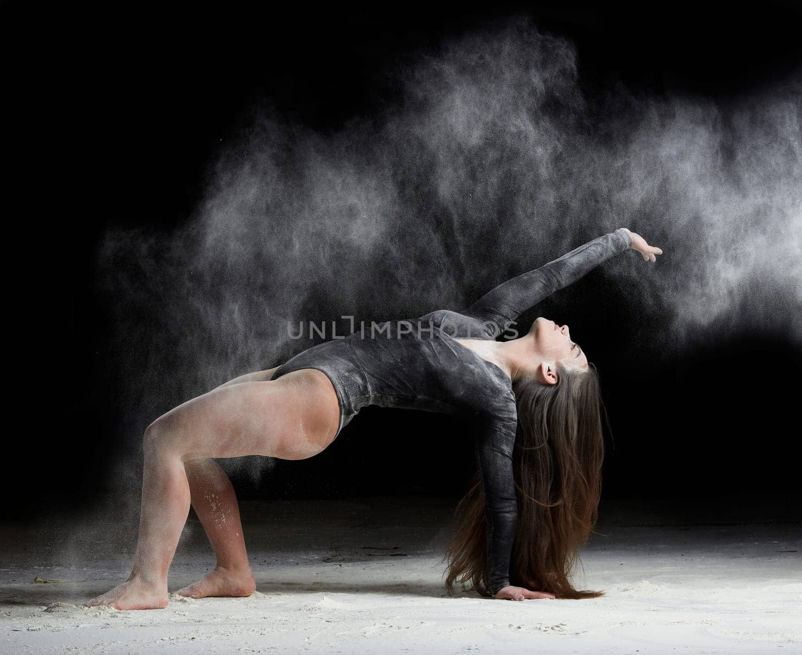 young beautiful caucasian woman with long hair is dressed in a sports black bodysuit stands in a bent pose and throws white flour, black background, gymnastic exercises
