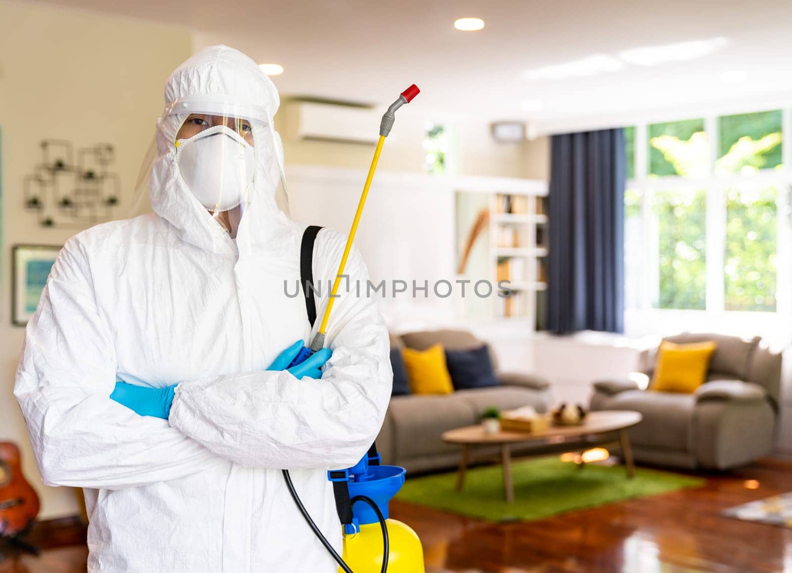 Portrait of medical staff in personal protective equipment PPE suit with coronavirus COVID-19 disinfectant spray with living room background. Delivery health care hygiene and disinfection concept