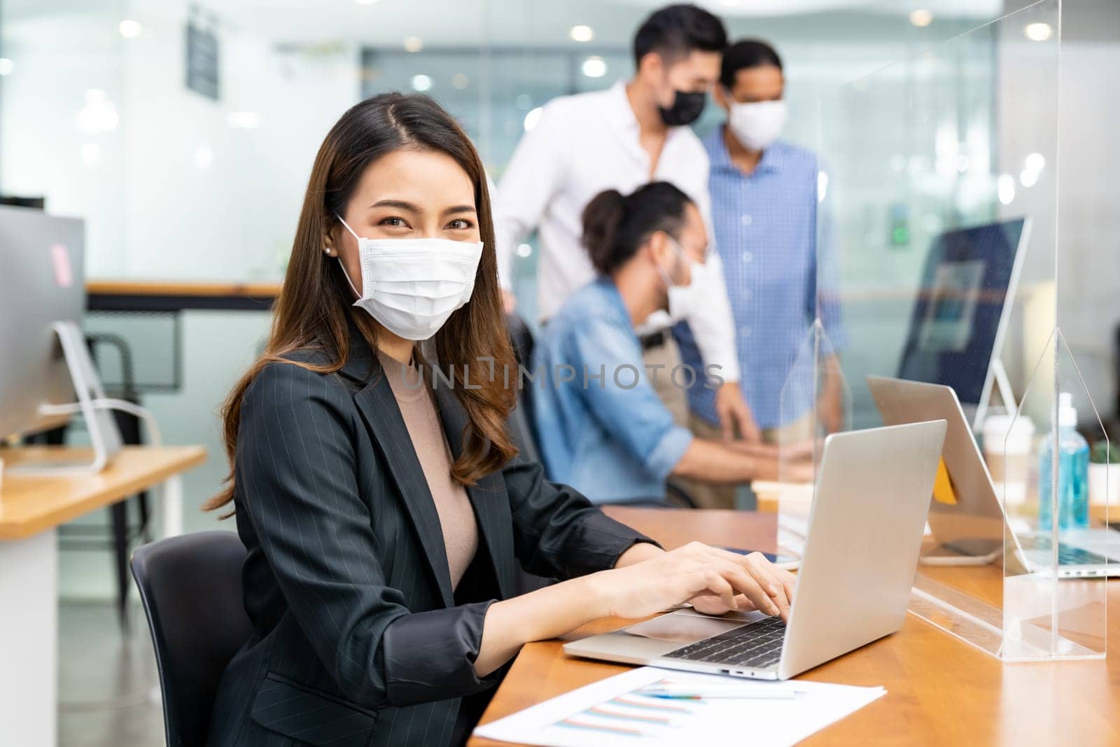 Portrait of asian office employee businesswoman wear protective face mask work in new normal office with interracial team in background as social distance practice prevent coronavirus COVID-19.