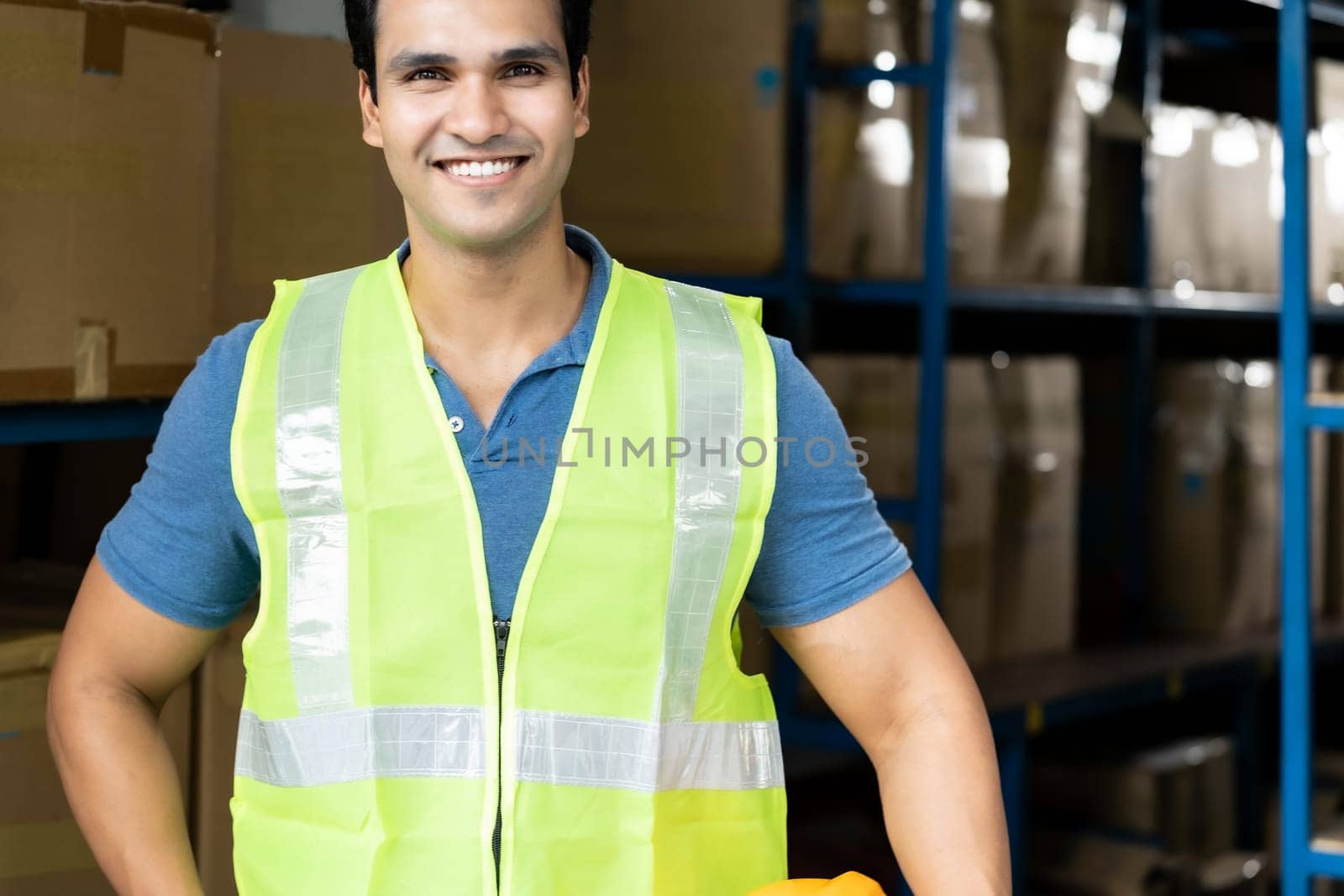 Portrait of Indian asian warehouse worker with safety vest stand and hold yellow helmet in warehouse distribution center environment. Using in business warehouse and logistic concept.