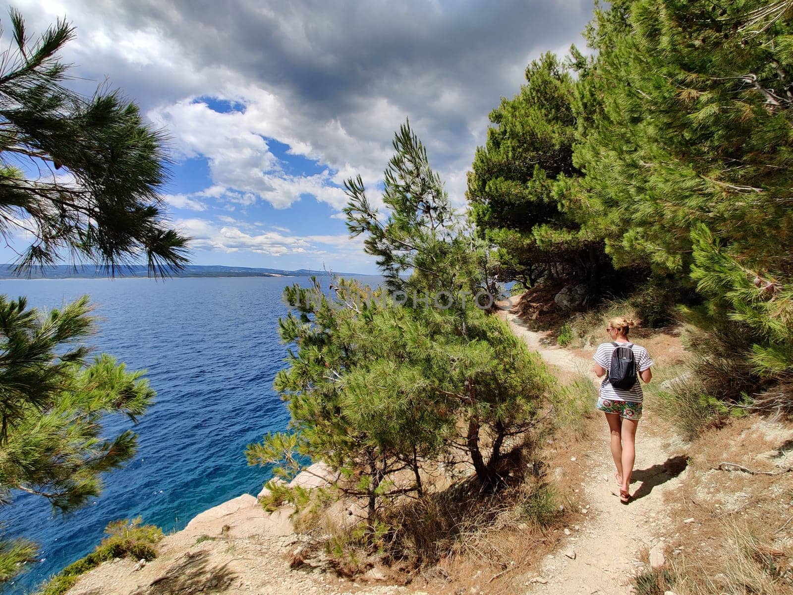 Young active feamle tourist wearing small backpack walking on coastal path among pine trees looking for remote cove to swim alone in peace on seaside in Croatia. Travel and adventure concept.