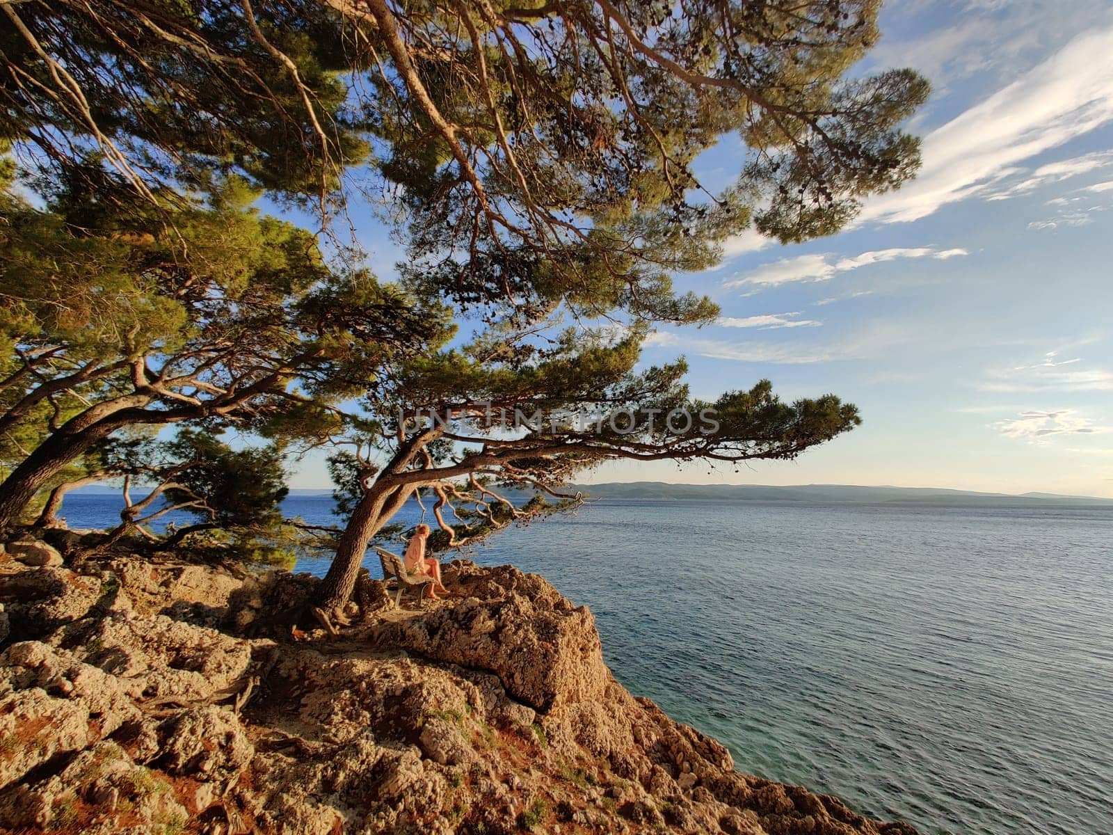 Pensive woman on vacations, sitting and relaxing under large pine tree on bench by dip blue sea enjoying beautiful sunset light in Brela, Makarska region, Dalmatia, Croatia.