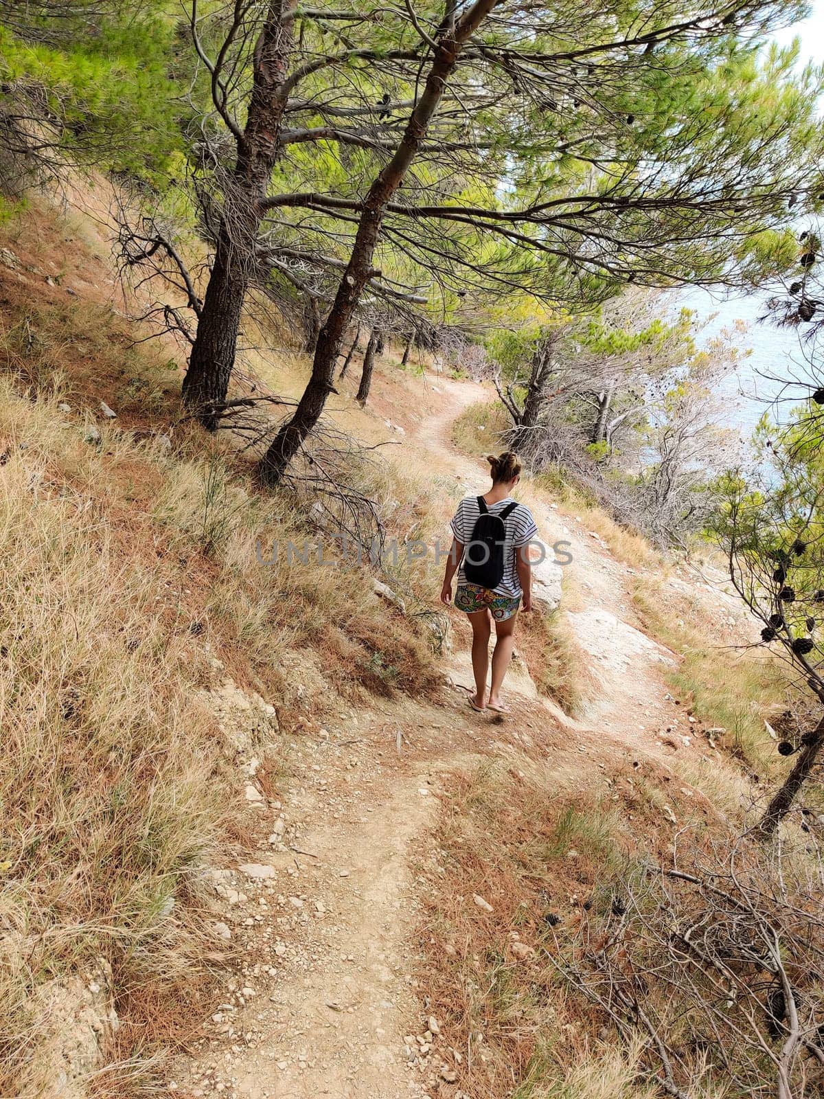 Young active feamle tourist wearing small backpack walking on coastal path among pine trees looking for remote cove to swim alone in peace on seaside in Croatia. Travel and adventure concept.
