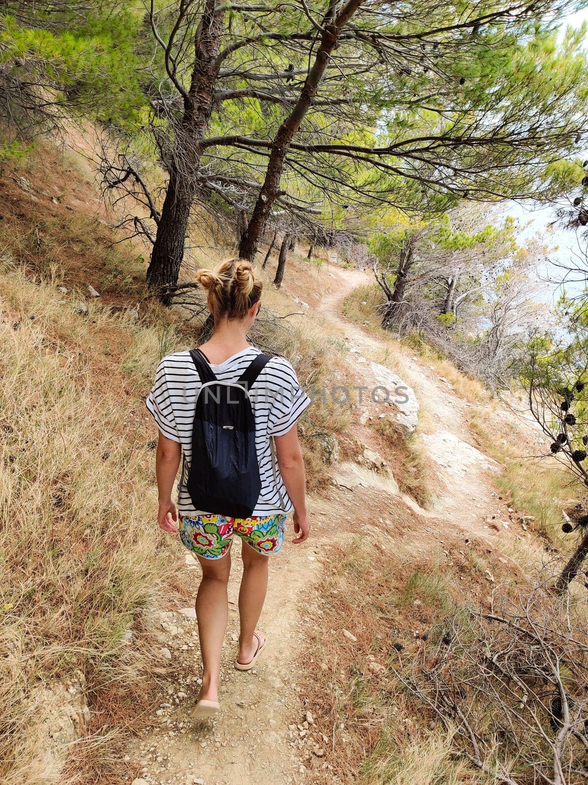 Young active feamle tourist wearing small backpack walking on coastal path among pine trees looking for remote cove to swim alone in peace on seaside in Croatia. Travel and adventure concept.