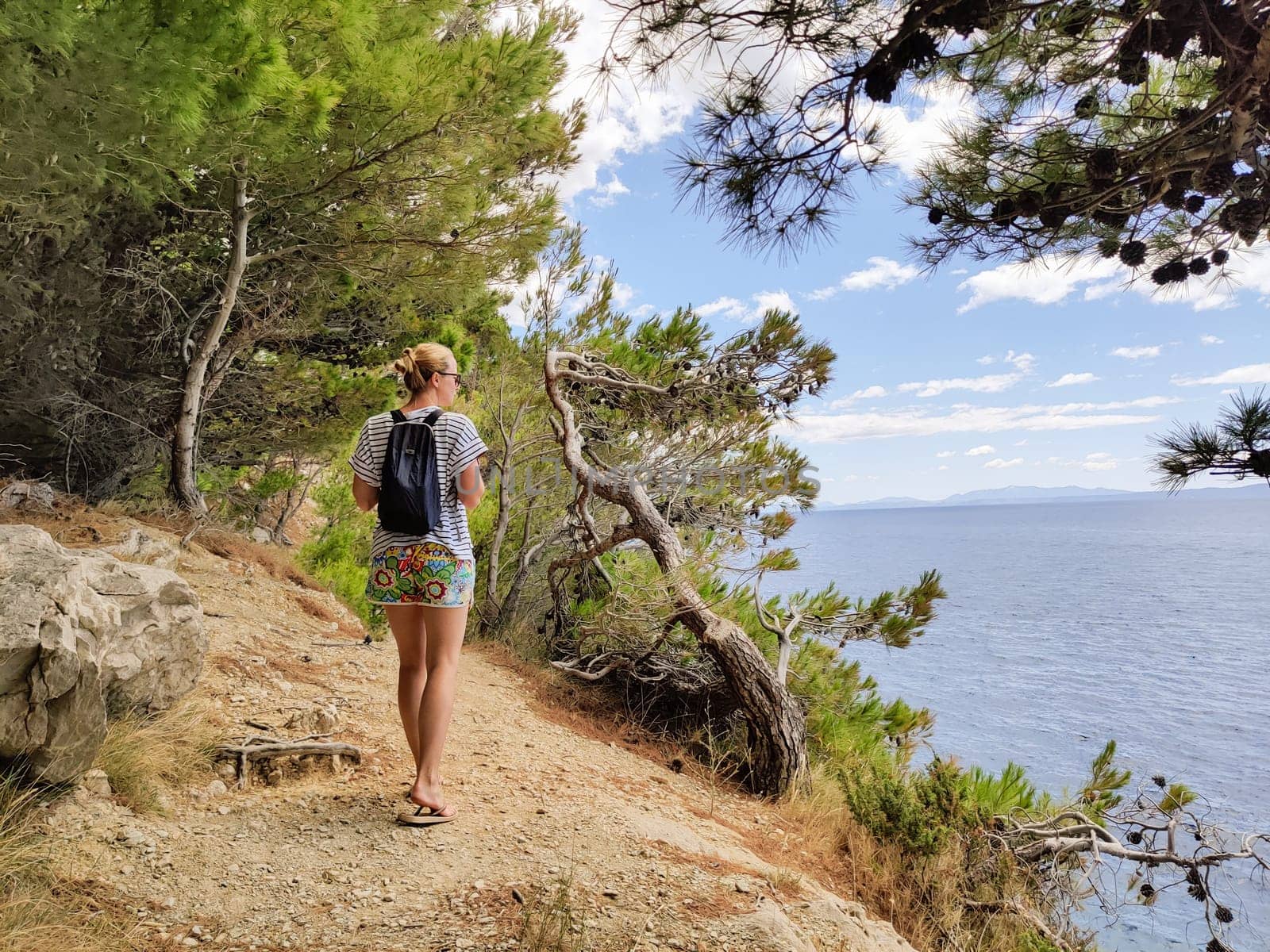Young active feamle tourist wearing small backpack walking on coastal path among pine trees looking for remote cove to swim alone in peace on seaside in Croatia. Travel and adventure concept.
