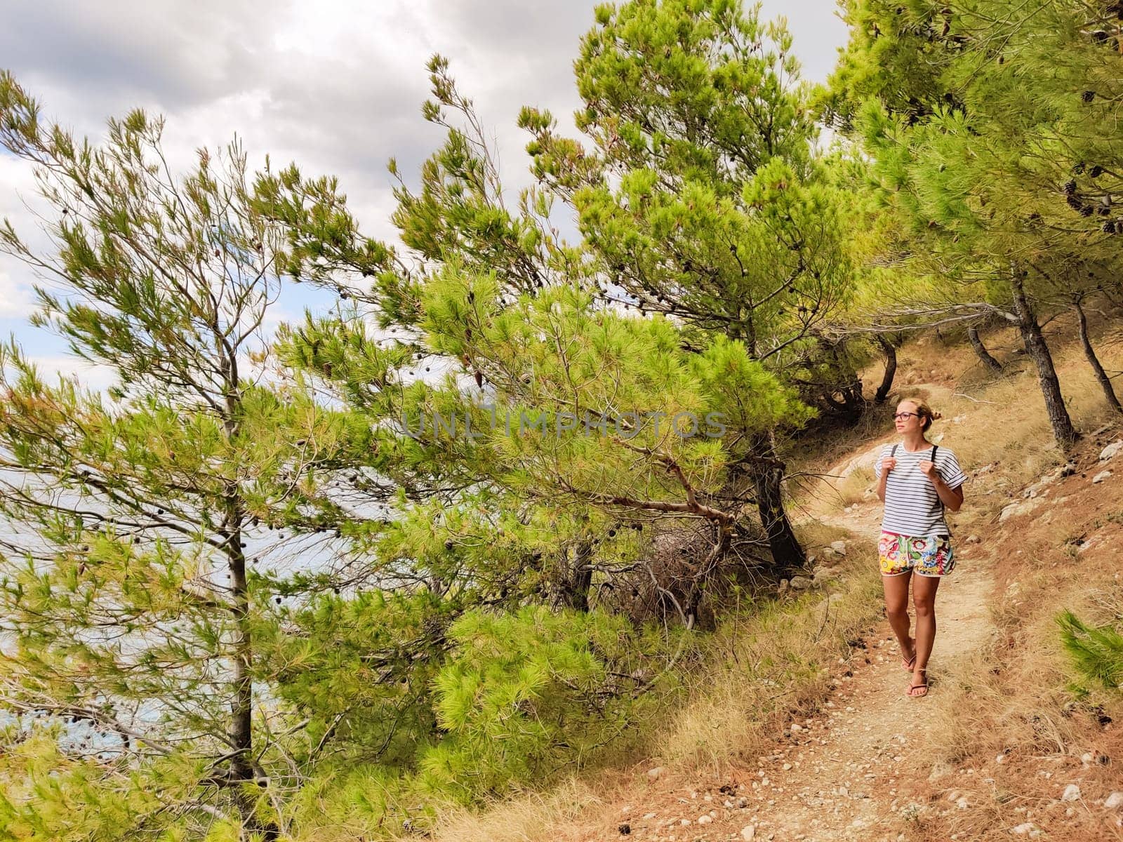 Young active feamle tourist wearing small backpack walking on coastal path among pine trees looking for remote cove to swim alone in peace on seaside in Croatia. Travel and adventure concept.