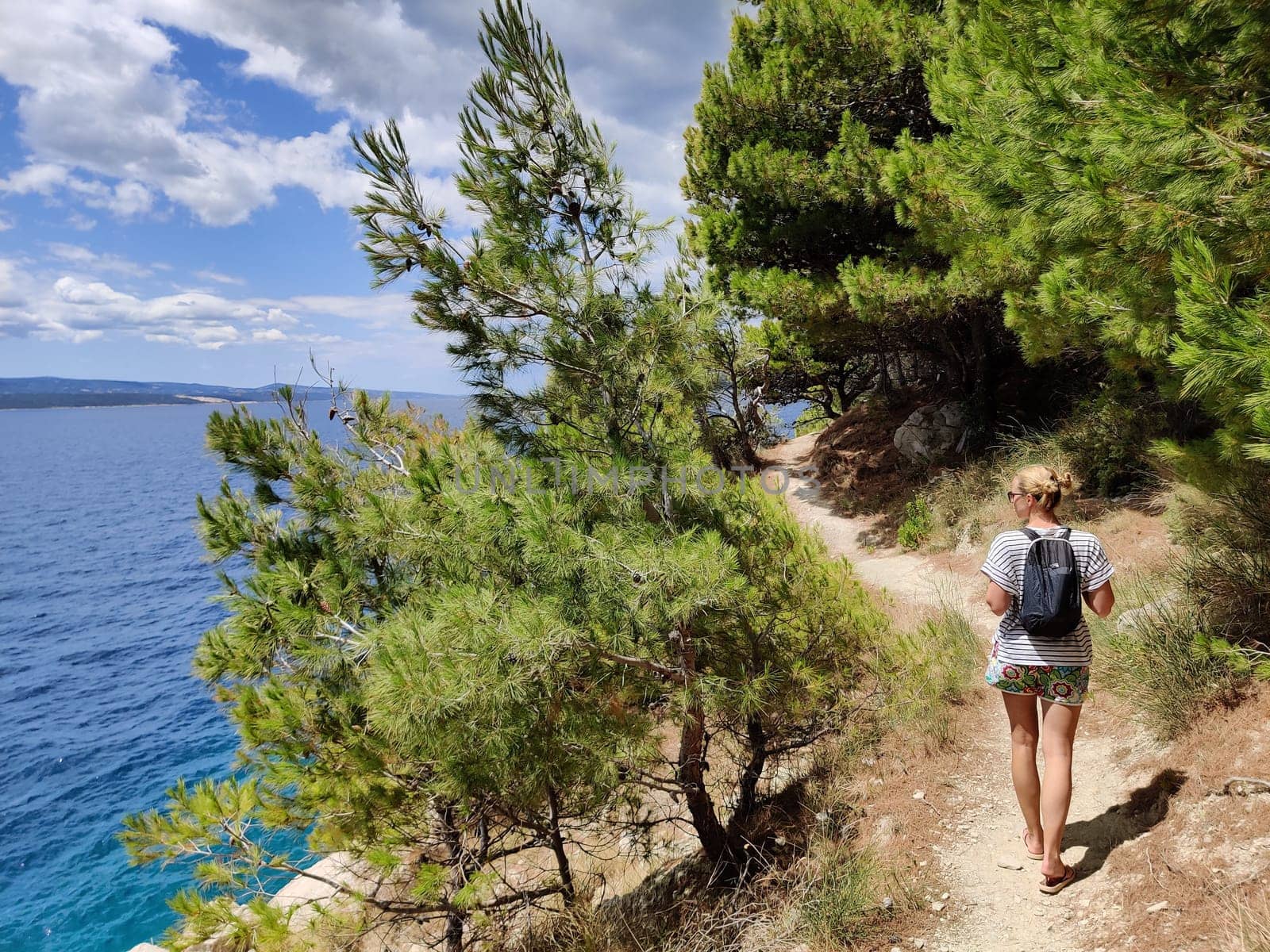Young active feamle tourist wearing small backpack walking on coastal path among pine trees looking for remote cove to swim alone in peace on seaside in Croatia. Travel and adventure concept.