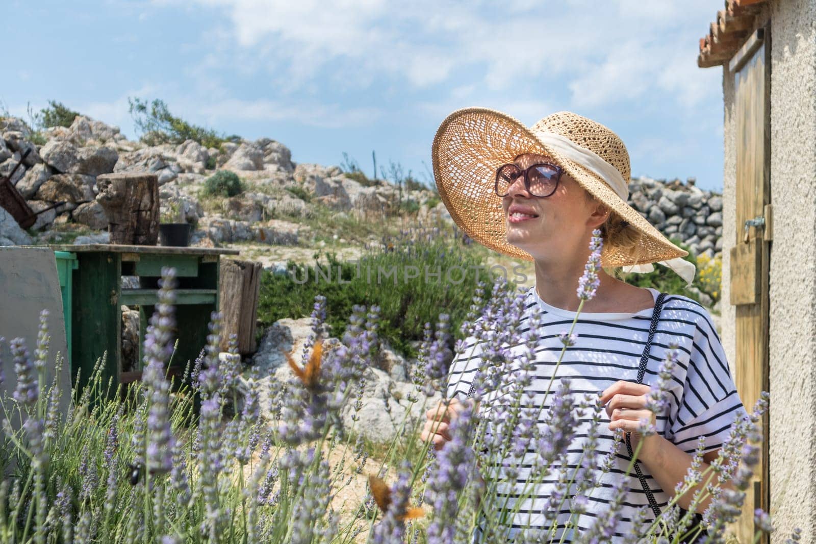 Beautiful blonde young female traveler wearing straw sun hat enjoying summer on Mediterranean cost strolling among lavander flowers on traditional costal village garden.