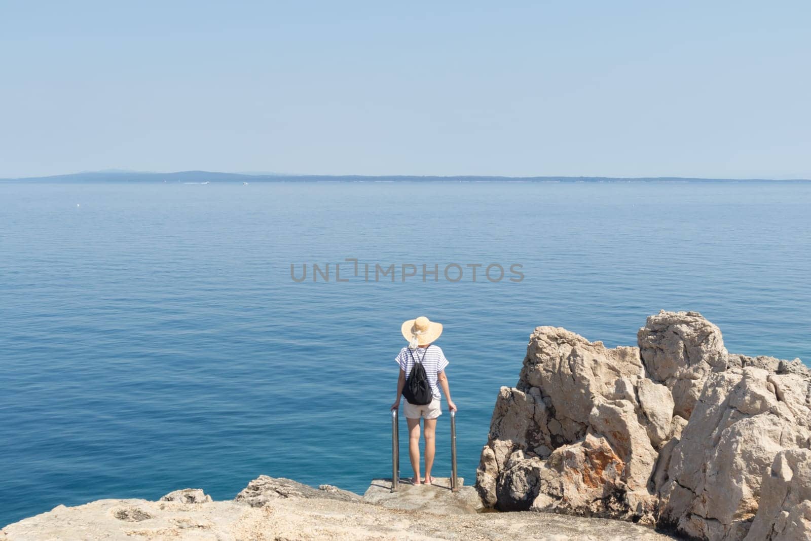 Woman traveler wearing straw summer hat and backpack, standing at edge of the rocky cliff looking at big blue sea and islands in on the horizon.