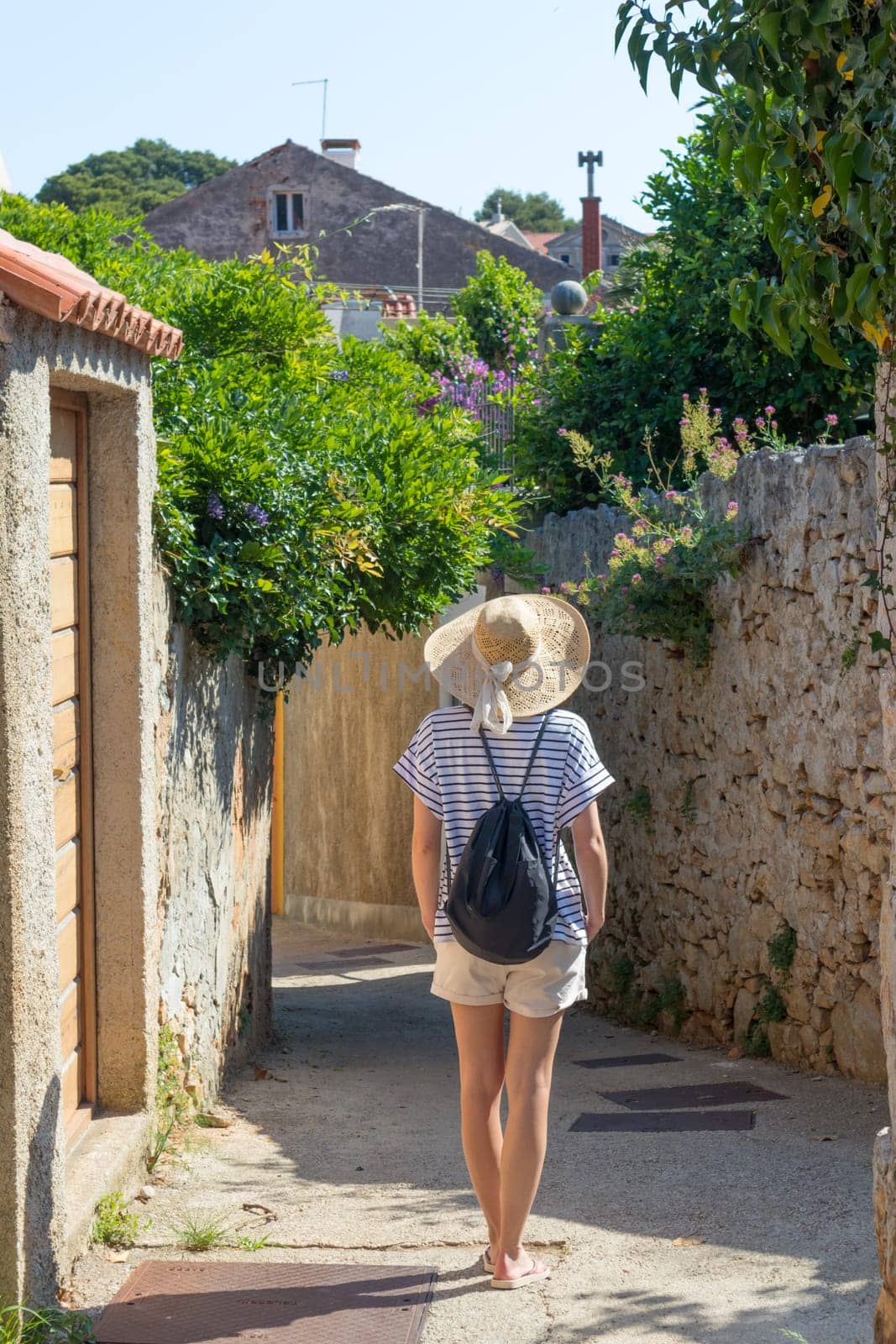 Rear view of beautiful blonde young female traveler wearing straw sun hat sightseeing and enjoying summer vacation in an old traditional costal town at Adriatic cost, Croatia.