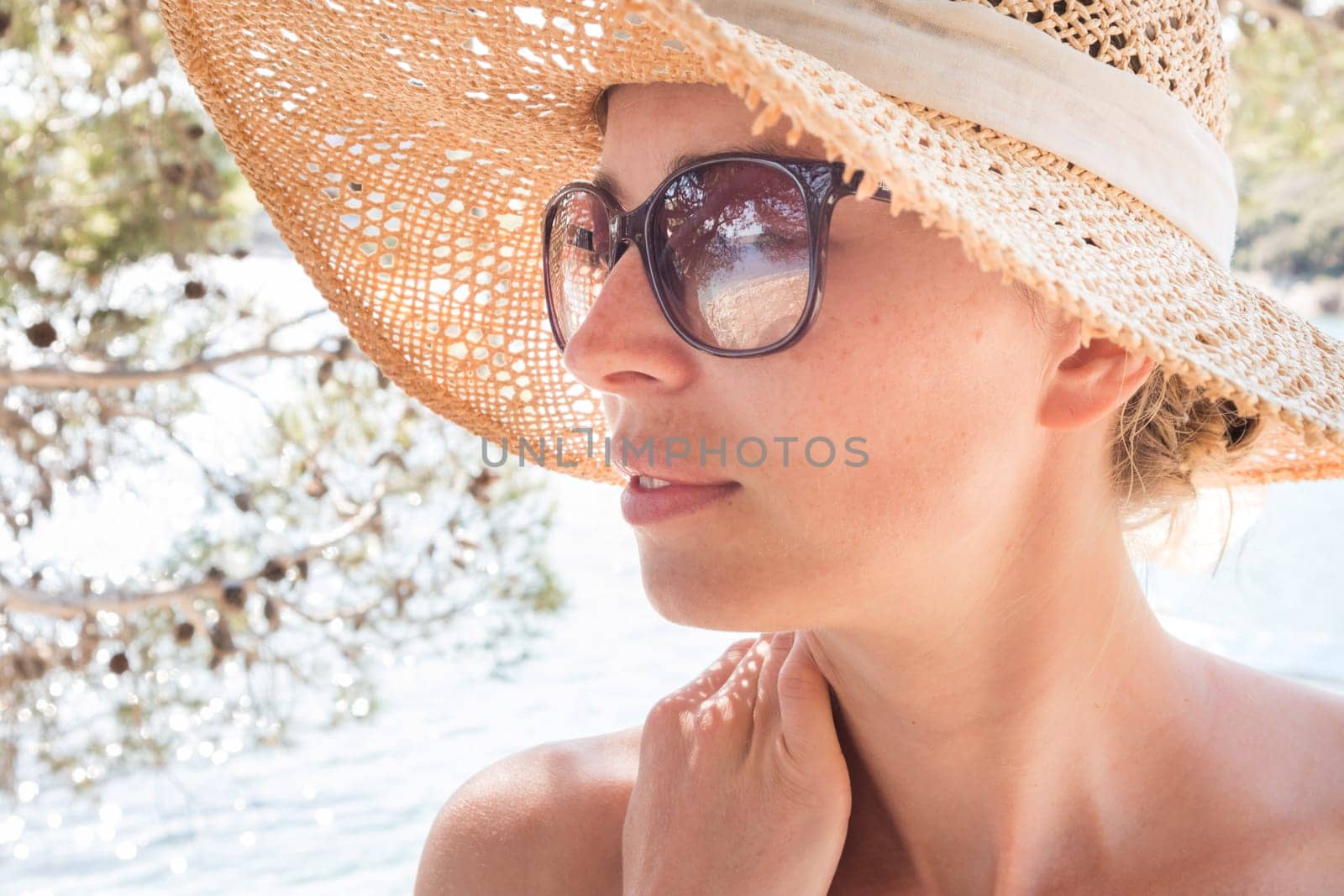 Close up portrait of no makeup natural beautiful woman wearing straw sun hat and sun glasses on the beach in shade of a pine tree.