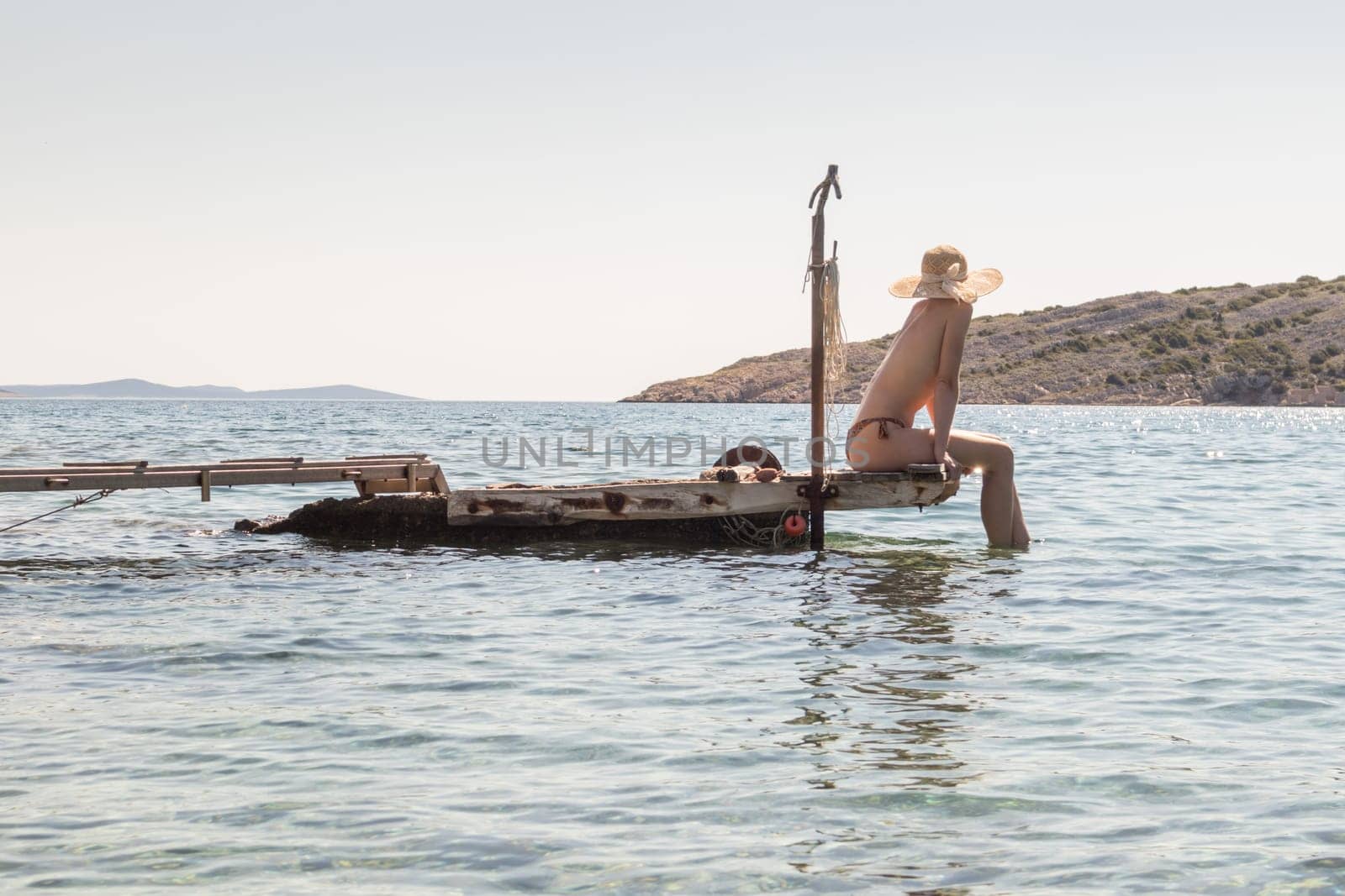 View of unrecognizable woman wearing big summer sun hat tanning topless and relaxing on old wooden pier in remote calm cove of Adriatic sea, Croatia.