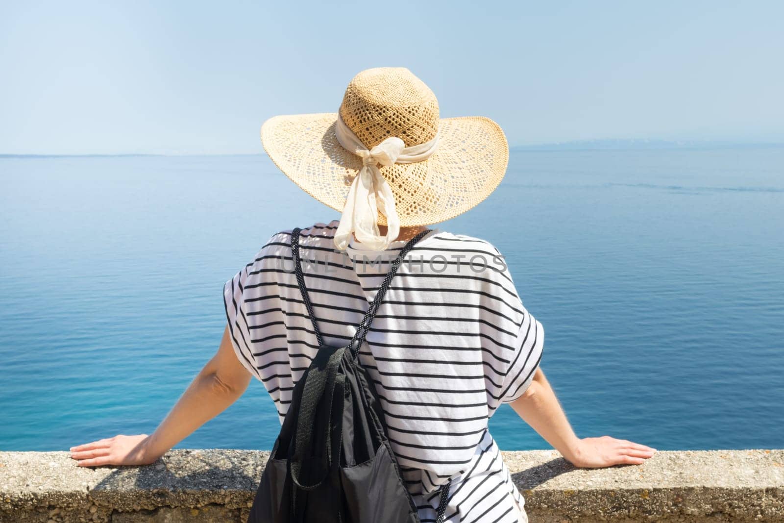 Rear view of woman traveler wearing straw summer hat and backpack,leaning against a stone wall looking at big blue sea and islands in on the horizon. Copy space.