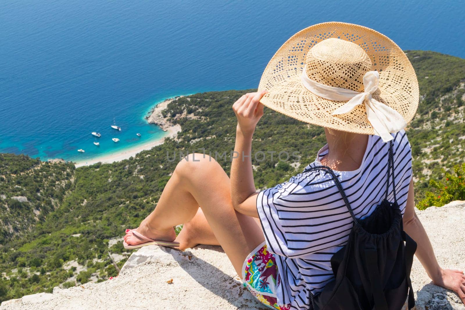 Active sporty woman on summer vacations sitting on old stone wall at Lubenice village, wearing straw hat and beach backpack enjoying beautiful coastal view of Cres island, Croatia.